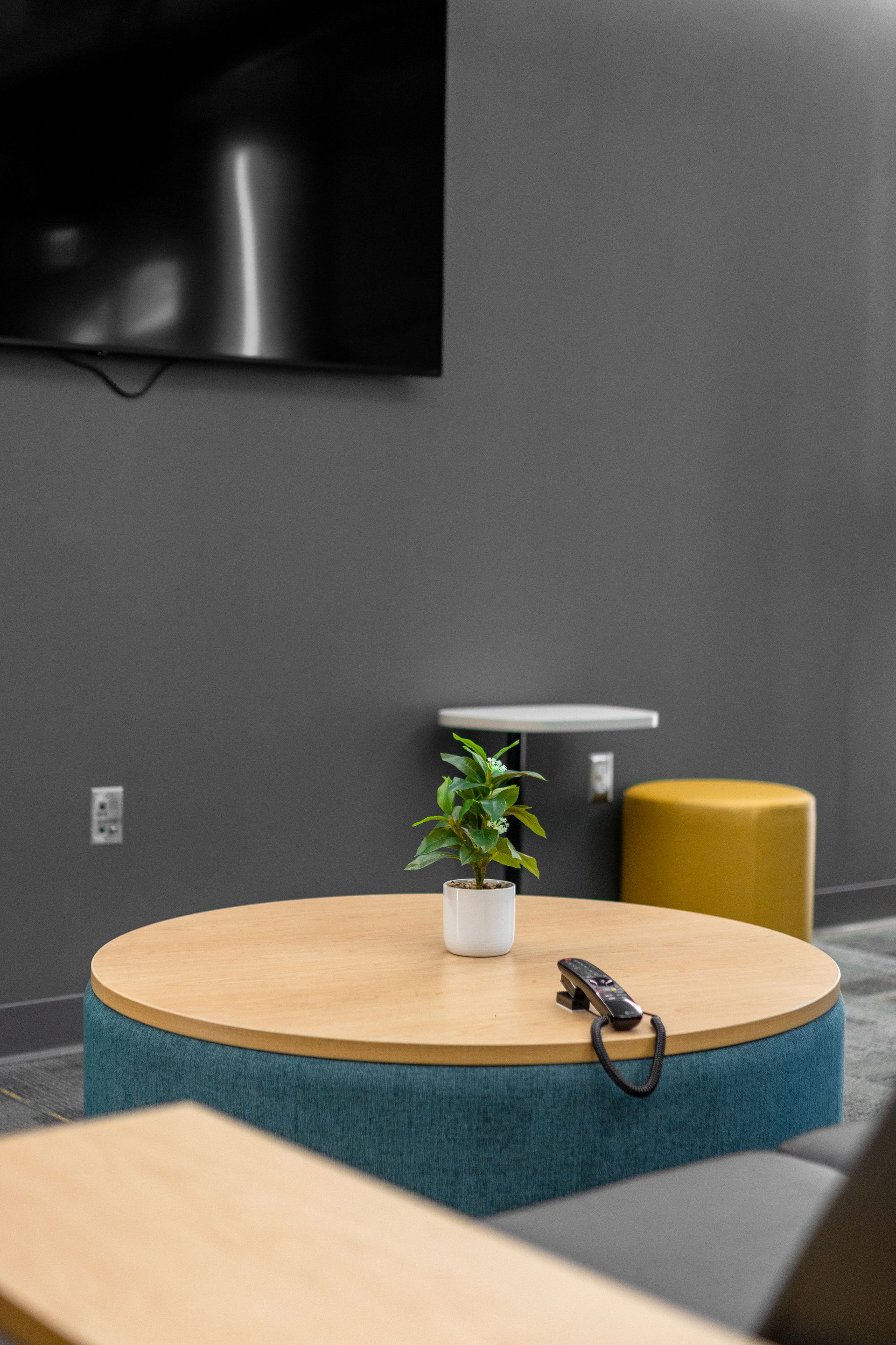 Eastview Terrace Kitchen's modern lounge with round wooden table, potted plant, headphones, and stools against a gray wall.