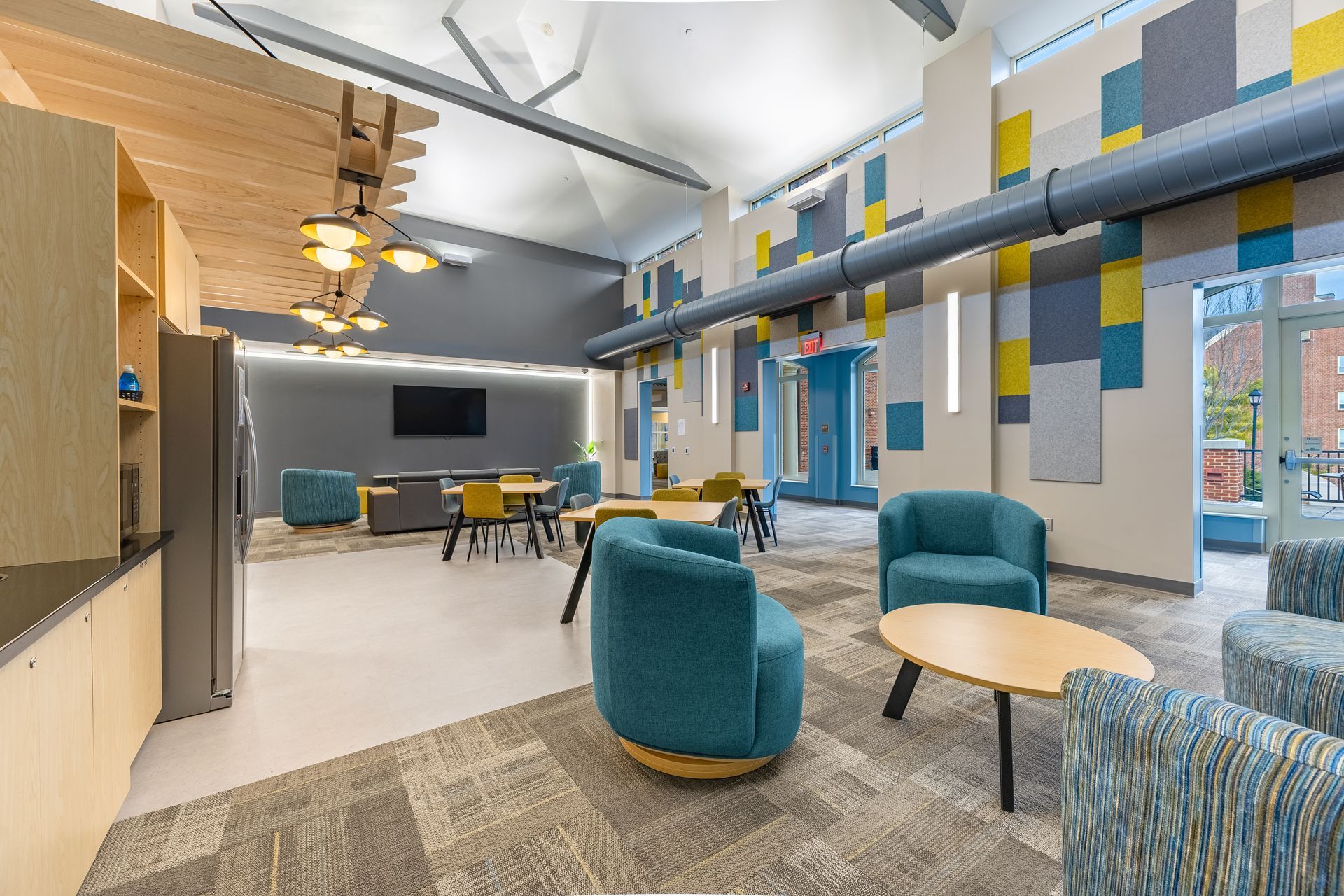 Eastview Terrace Kitchen's modern lounge with teal chairs, patterned rug, round tables, and colorful wall panels under high ceilings