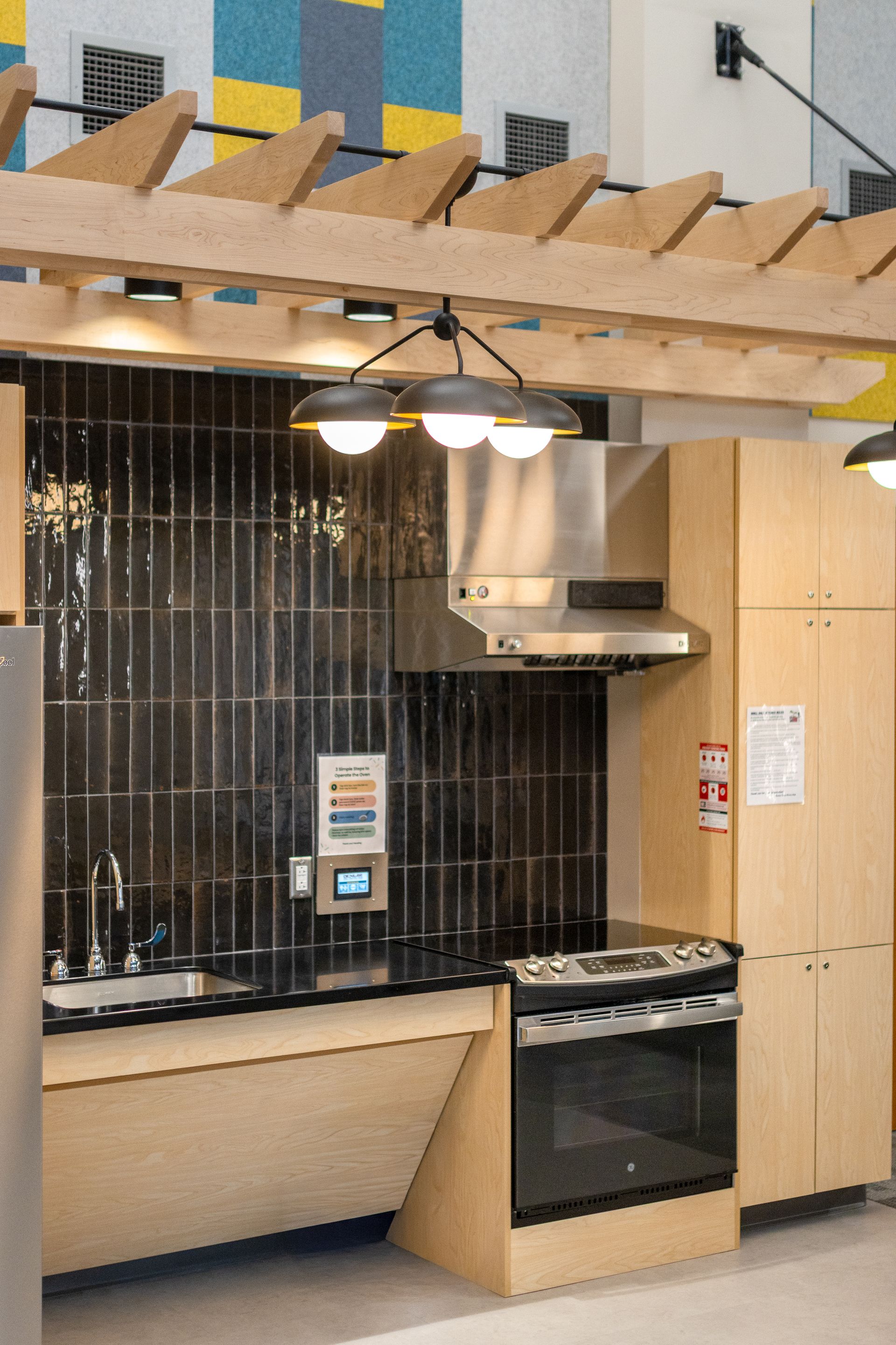Eastview Terrace Kitchen with black tile backsplash, beige cabinets, stove, and hanging pendant lights under a wood pergola