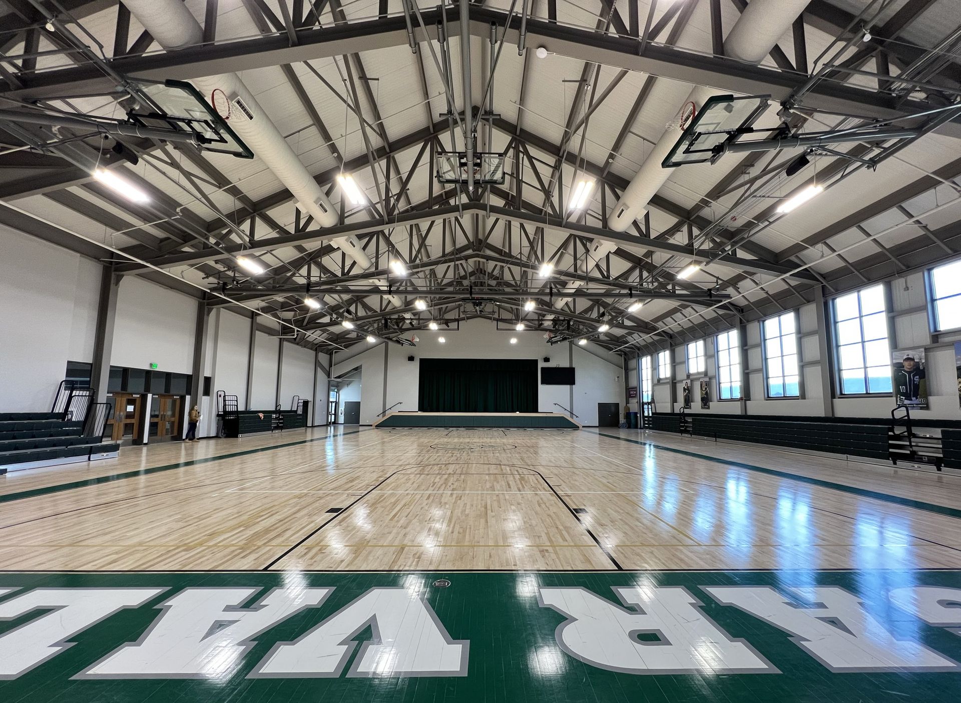 Wide-angle view of a gymnasium with a hardwood basketball court, exposed metal ceiling trusses, and bleachers.