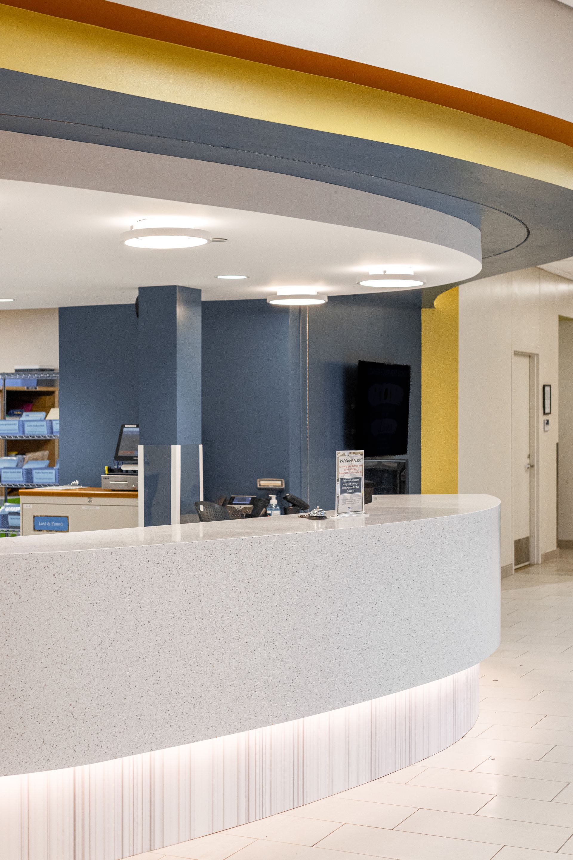 Reception desk with a curved white counter and blue tiled wall. Overhead, a curved ceiling design with yellow and gray accents.