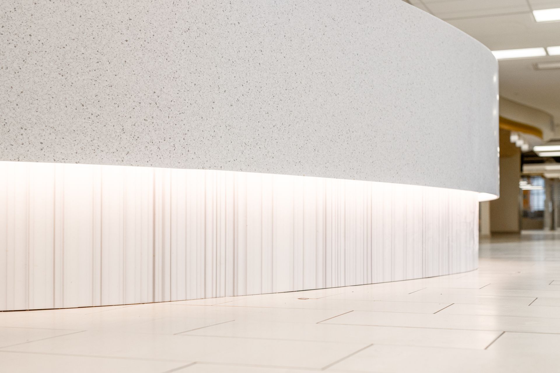 White, curved counter with textured surface, illuminated base, in a hallway with white tile floor.
