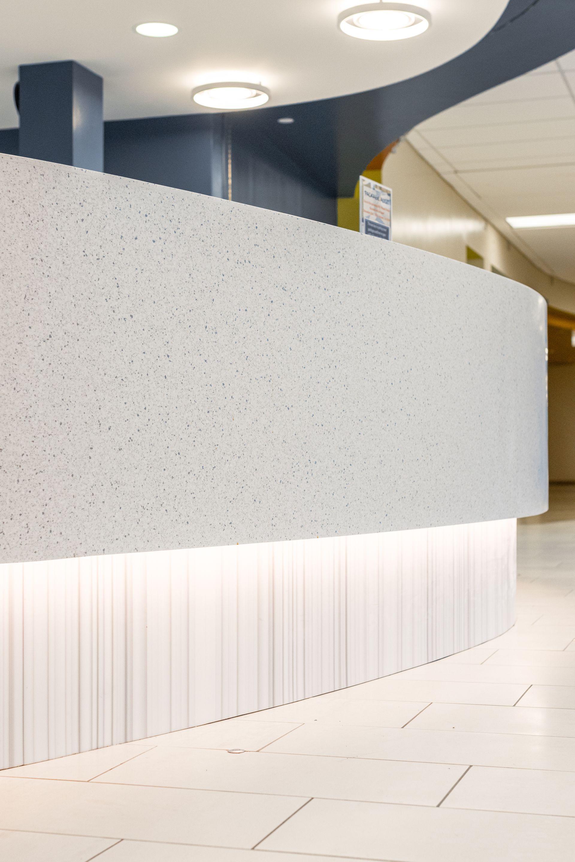 Curved, speckled white reception desk with vertical paneling, under bright circular lights, in a modern, light-filled space.