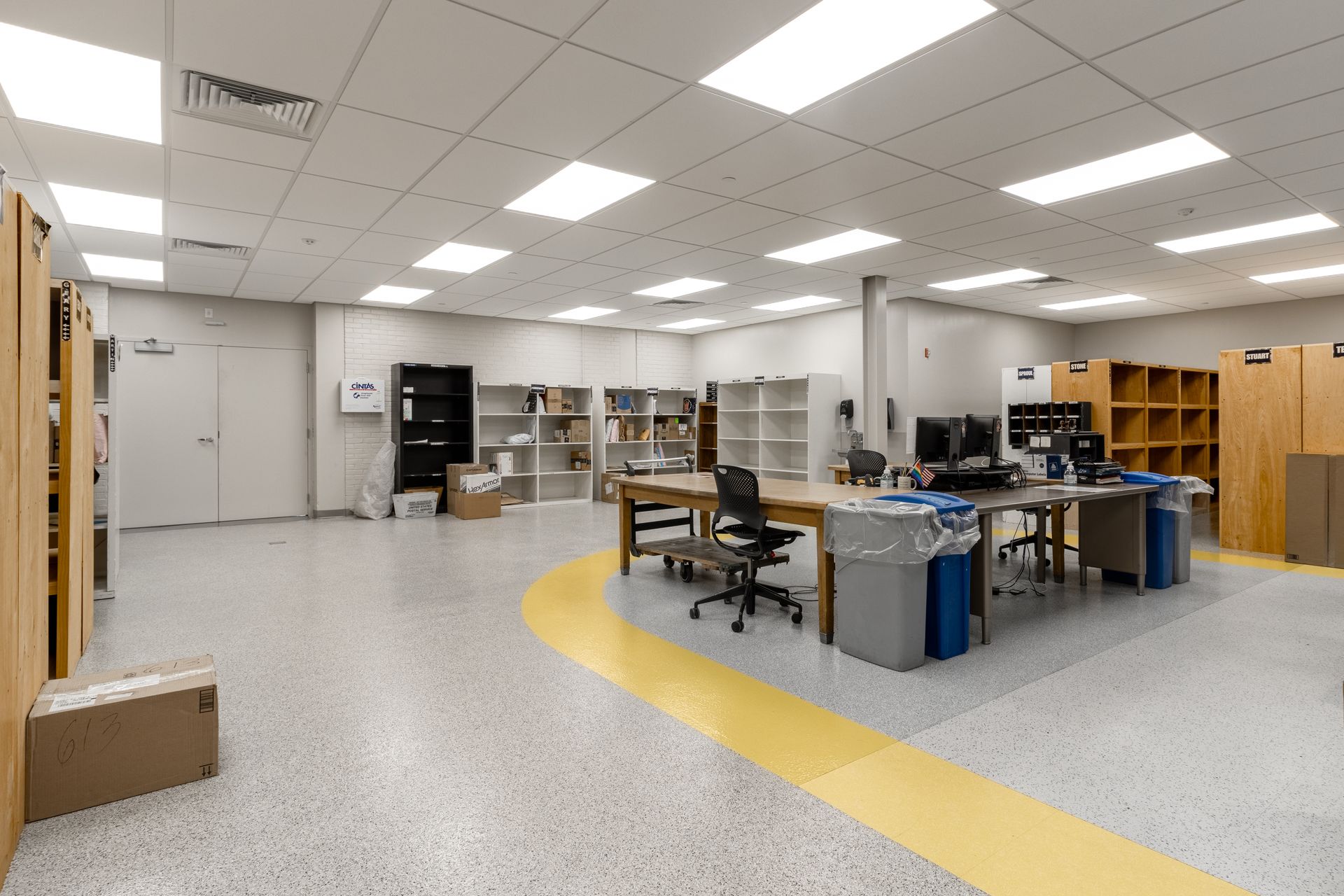 Interior of a brightly lit office with desks, computers, shelves, and a yellow pathway on the speckled floor.
