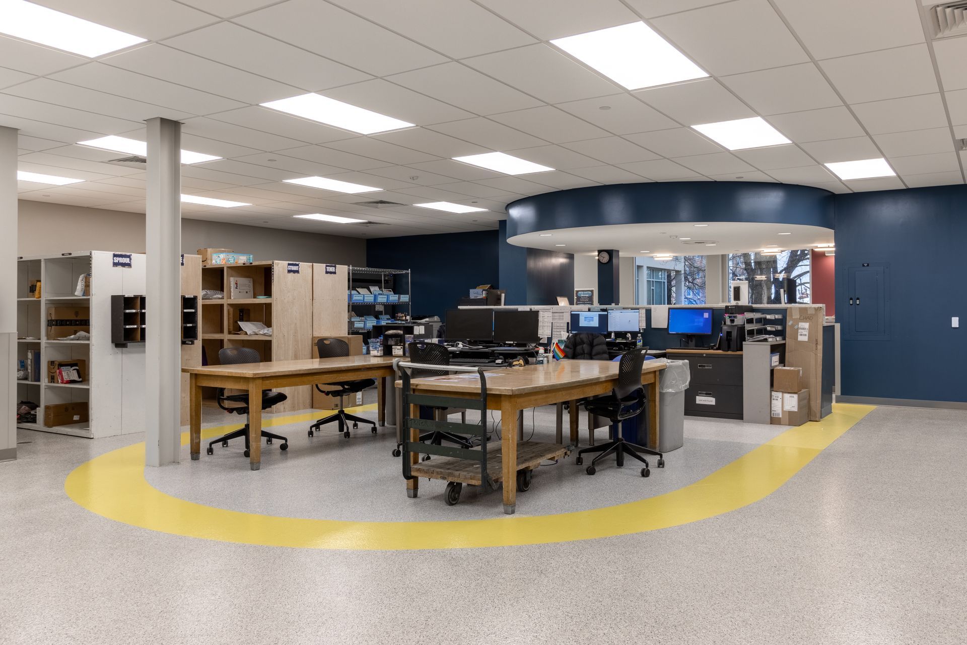 Open-plan office with desks, computers, and shelving. A yellow path guides through the space.