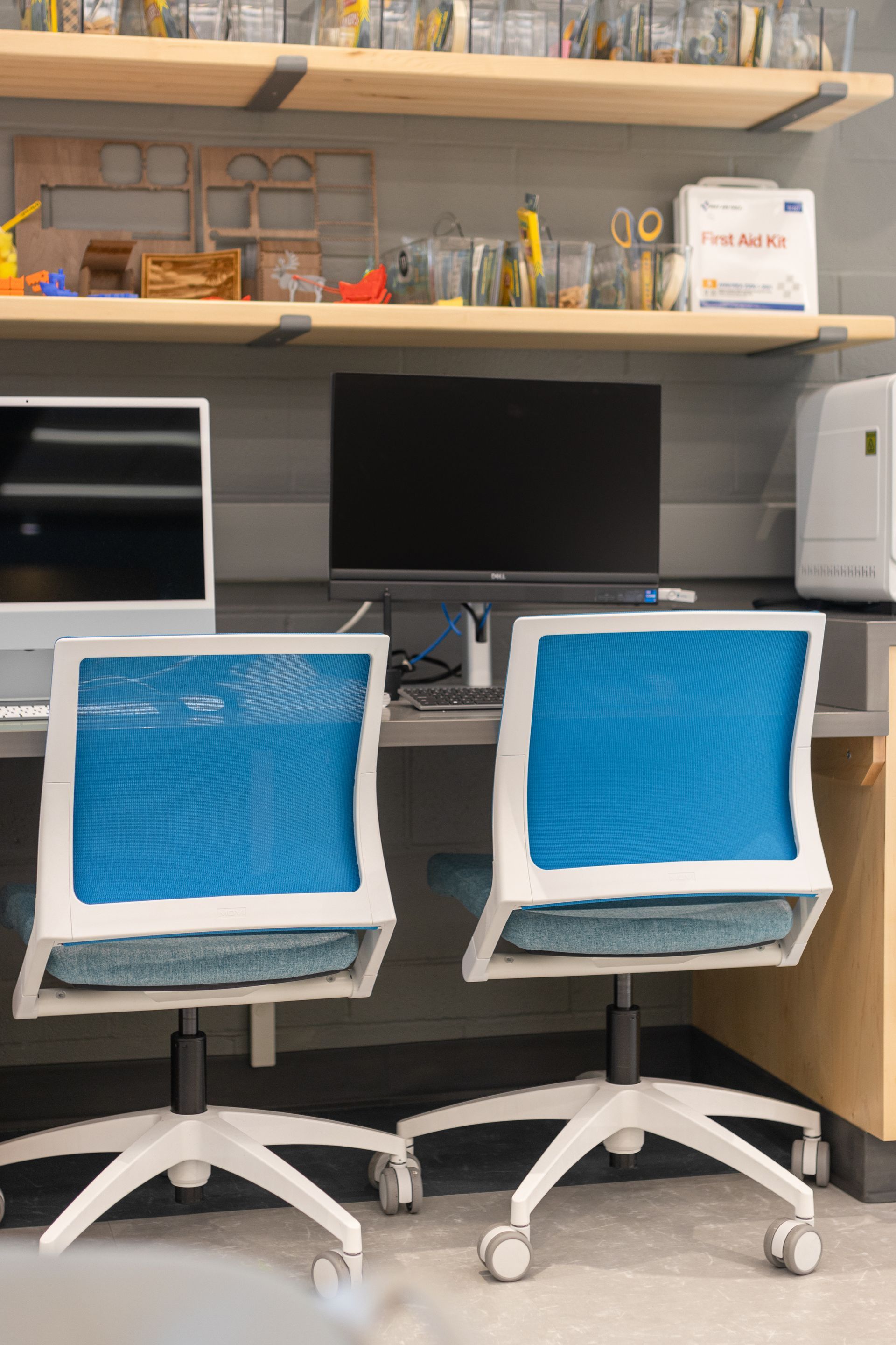 Two blue mesh office chairs at a desk with two computer monitors and shelves of supplies.