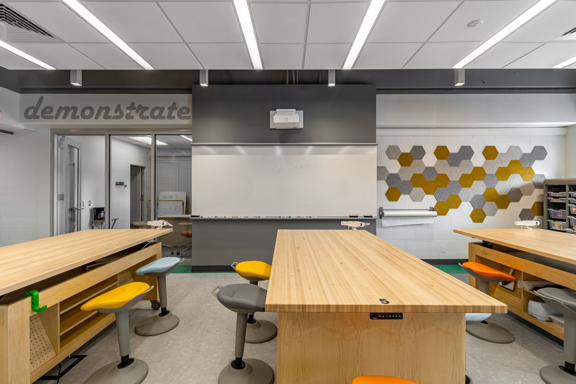 Modern classroom with wooden work tables, colorful stools, whiteboard, and honeycomb wall design.