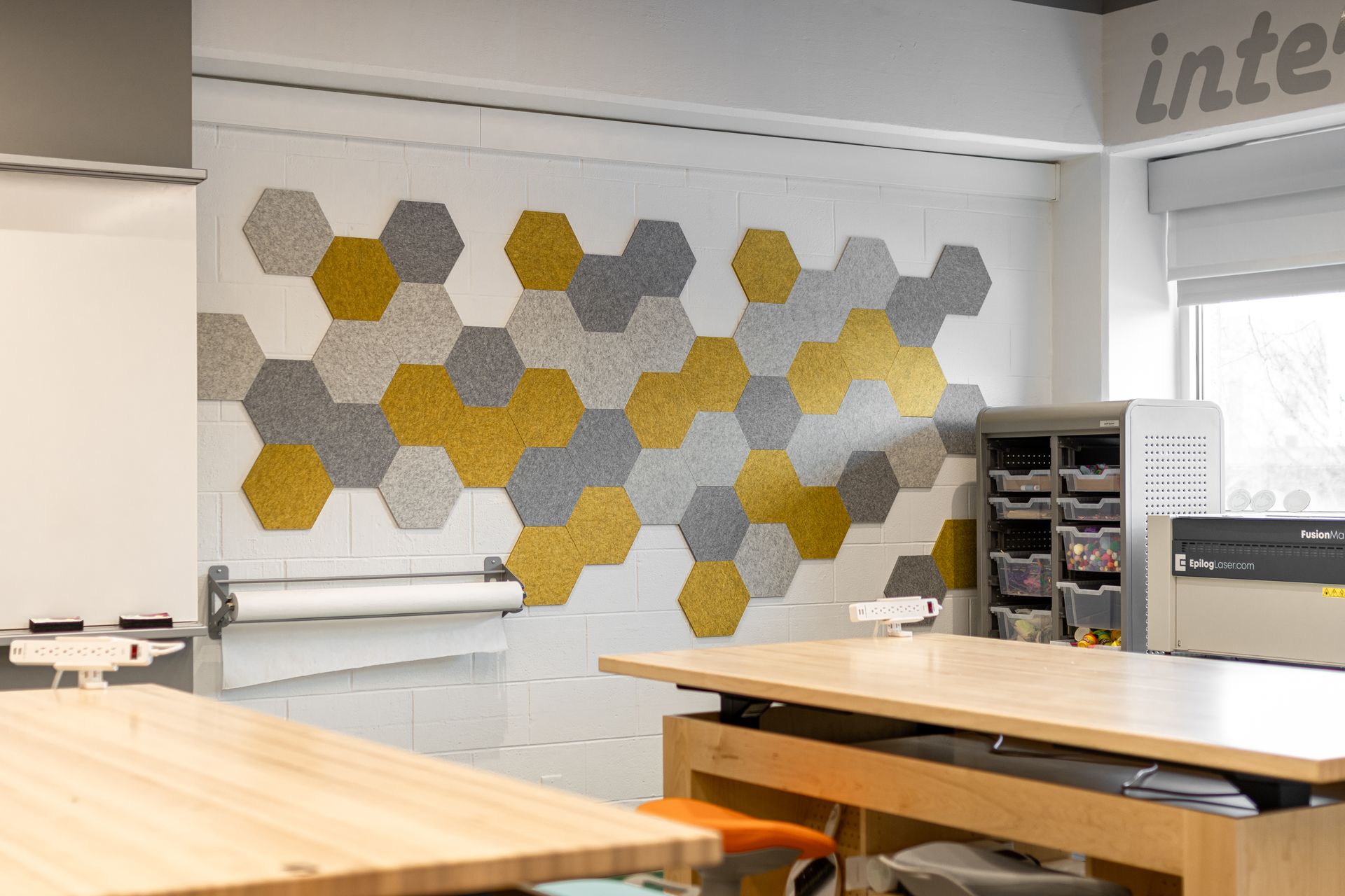 Hexagon-tiled wall with gray and yellow accents in a workshop. Tables and equipment are in the foreground.