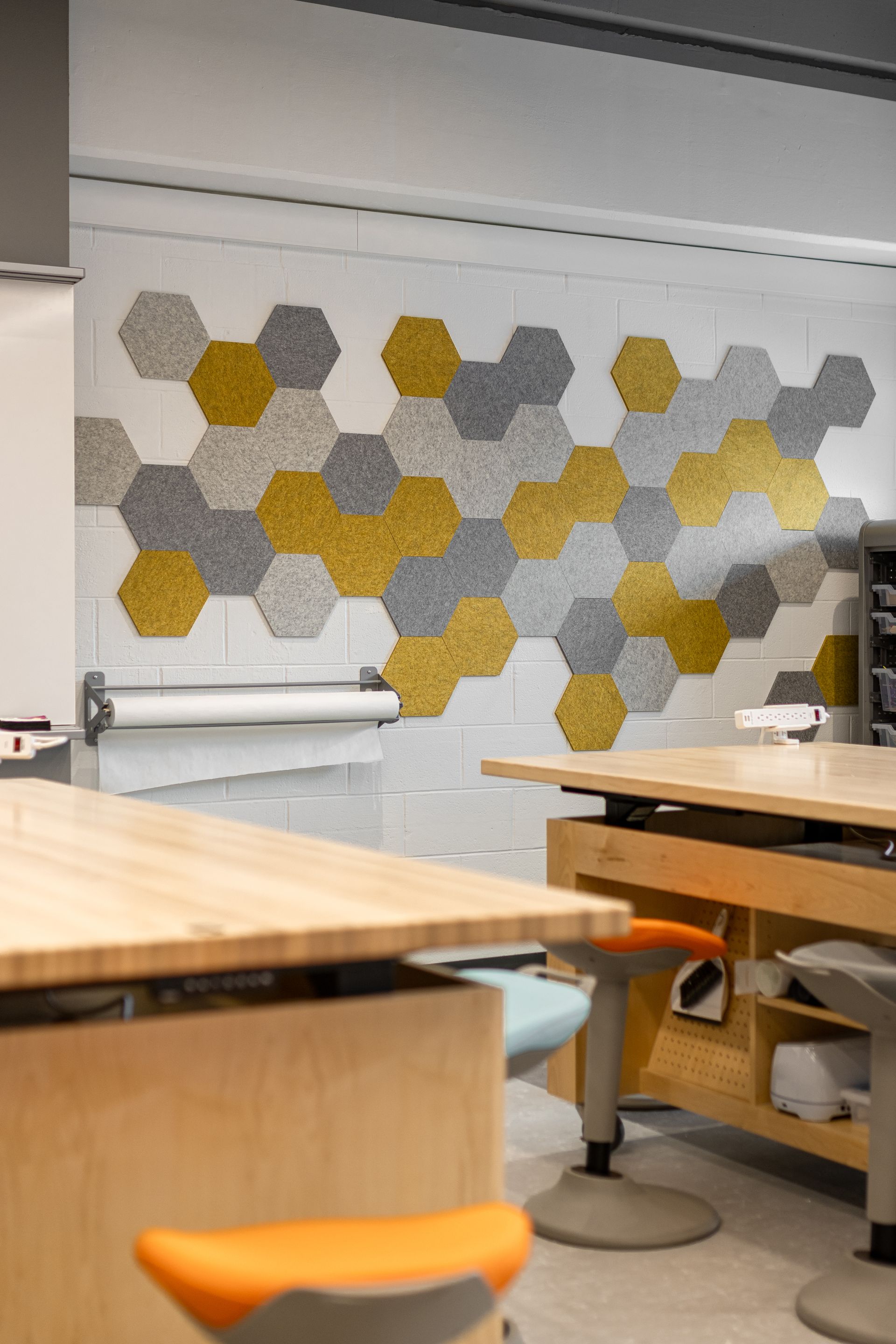 Classroom wall with gray, yellow, and white honeycomb-shaped acoustic panels, with wooden work tables and stools.