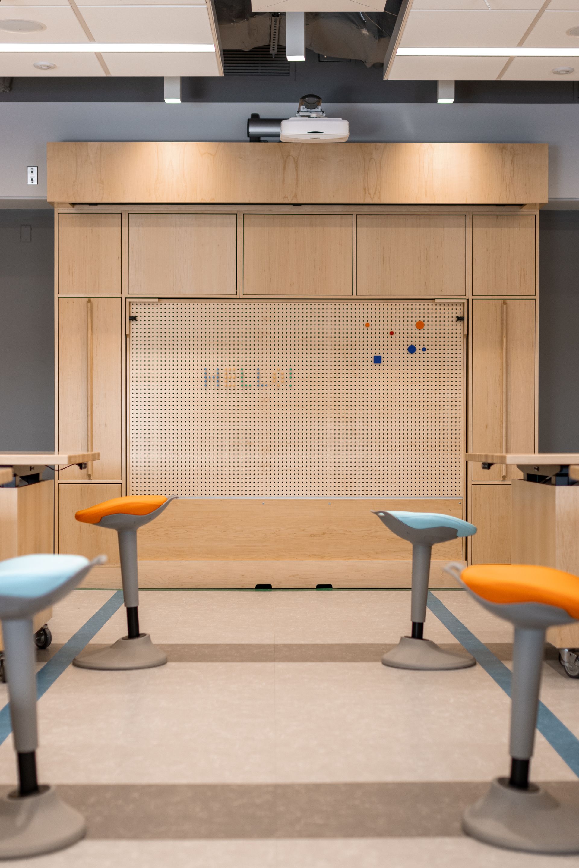 A classroom with a wooden pegboard, stools with blue and orange seats, and a projector.
