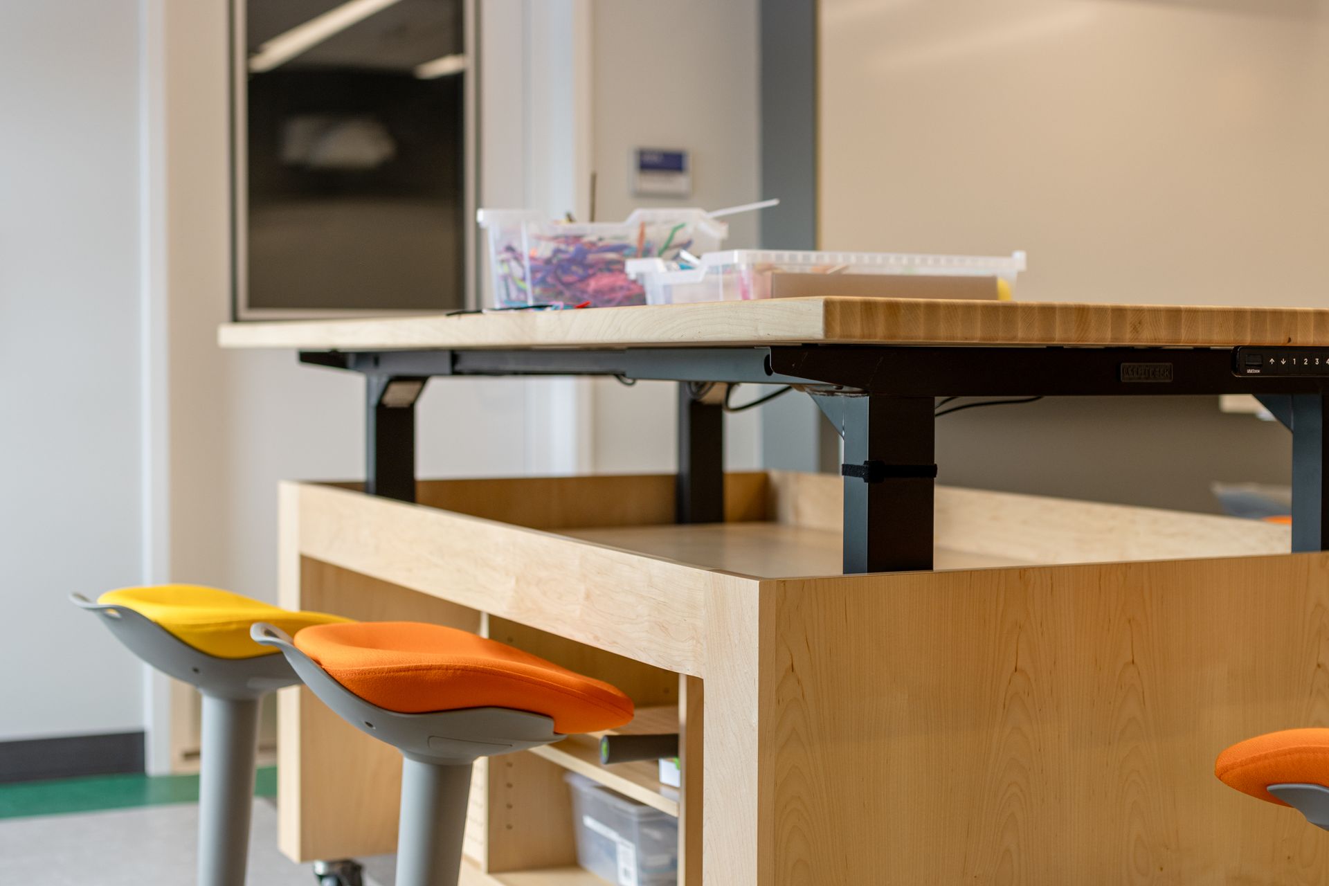 Height-adjustable wooden table with supplies, yellow and orange stools.