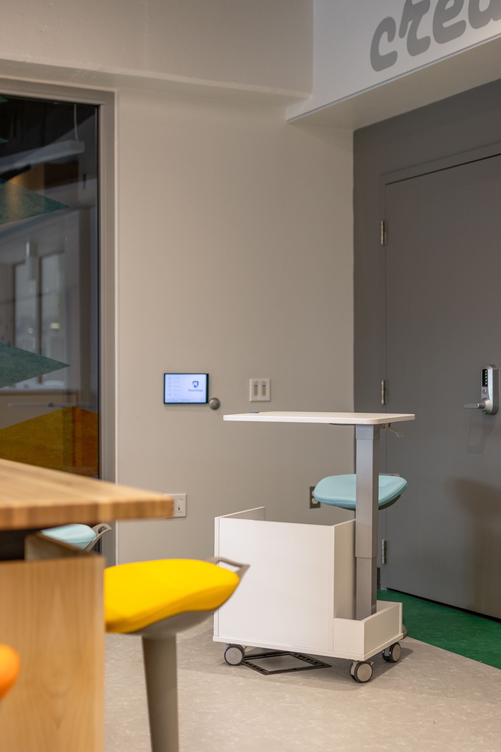 A mobile white table with a blue seat in a room, near a yellow stool and a gray door.