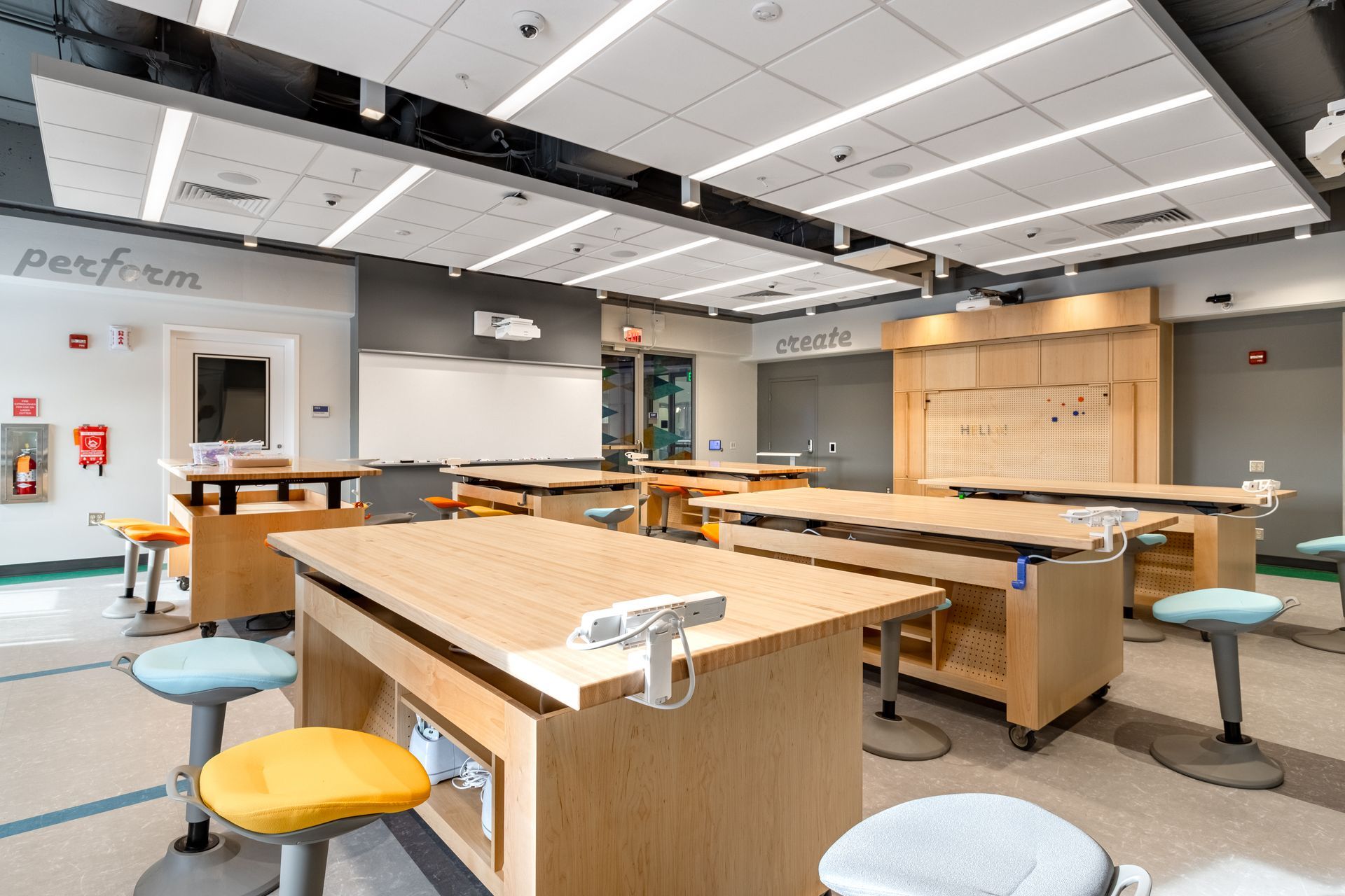A modern classroom with wooden work tables and adjustable stools. Bright fluorescent lighting.