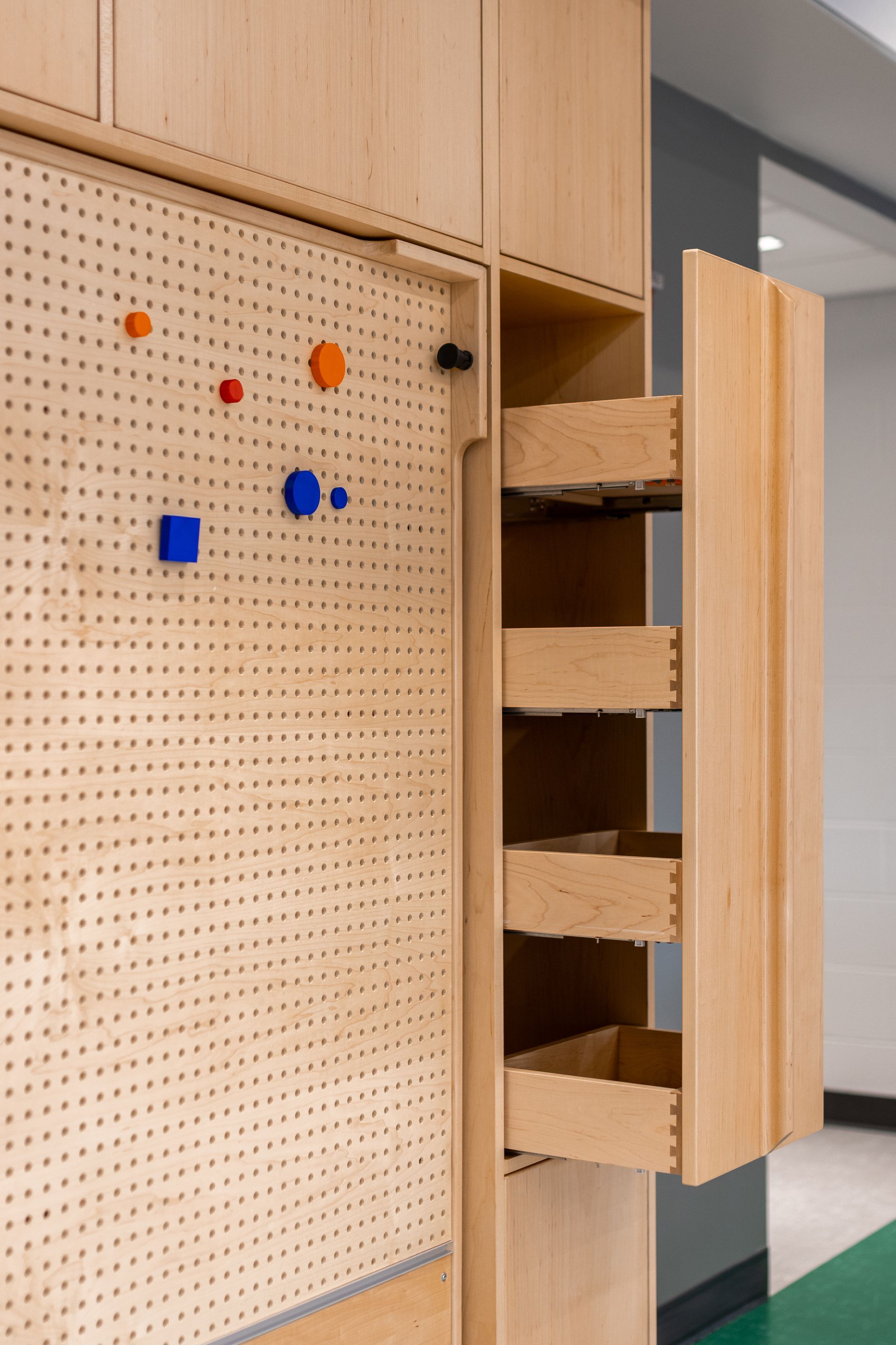 Light wood storage cabinet with open drawers and pegboard with colorful, geometric shapes attached.