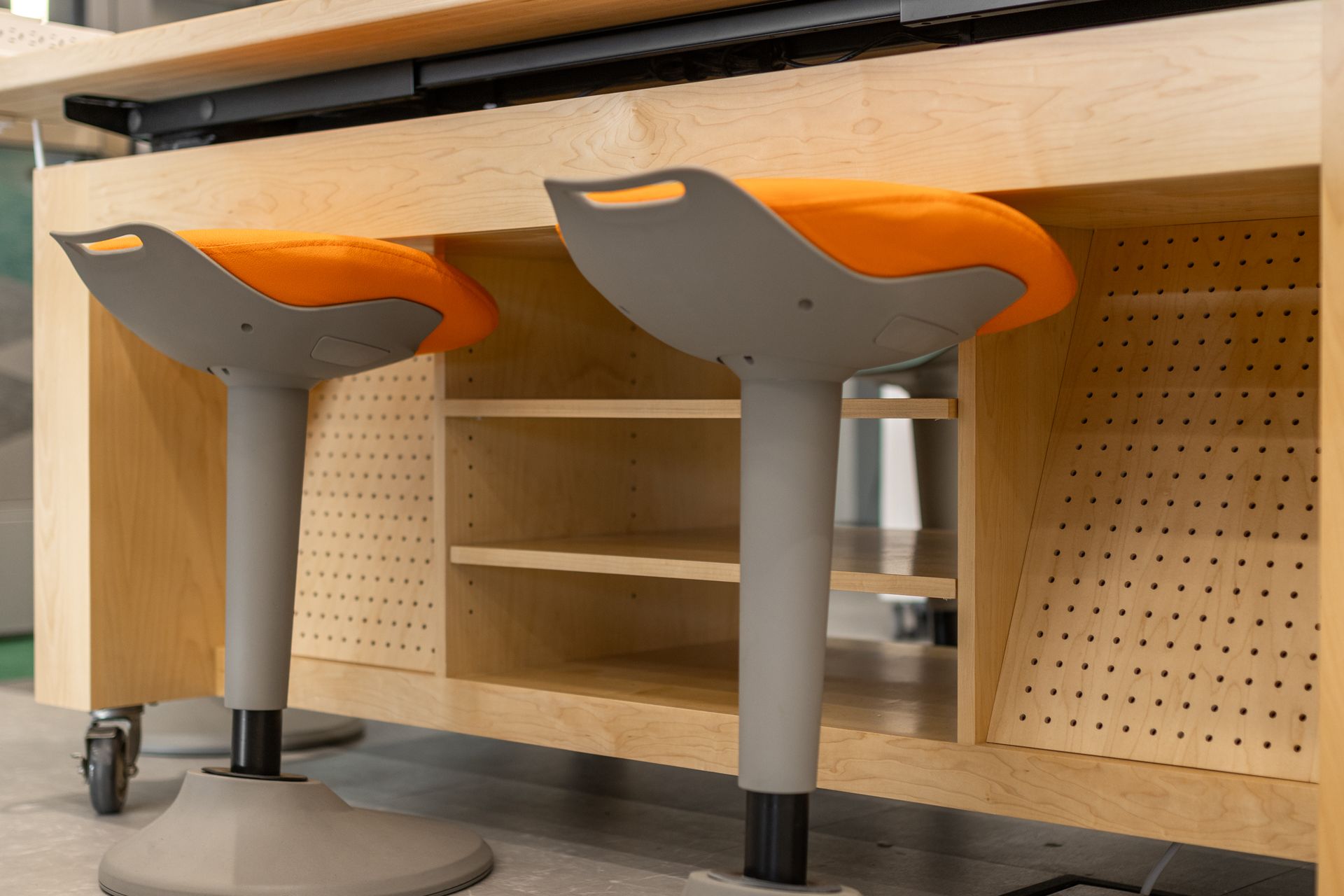 Two orange and gray stools sit at a light wood workstation with shelving and a black bar above.