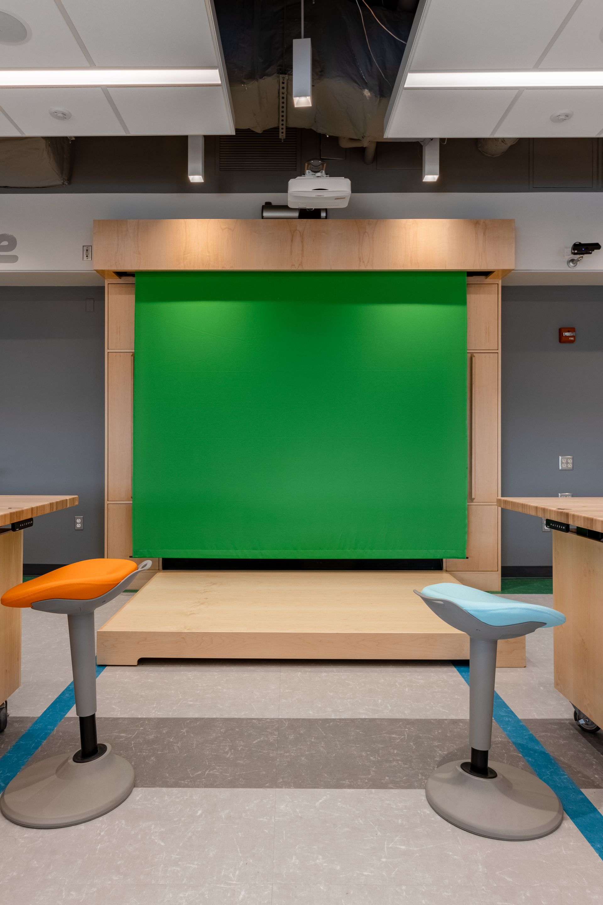 Green screen in a studio-like classroom setting, with two adjustable stools in front of it and workstations on either side.