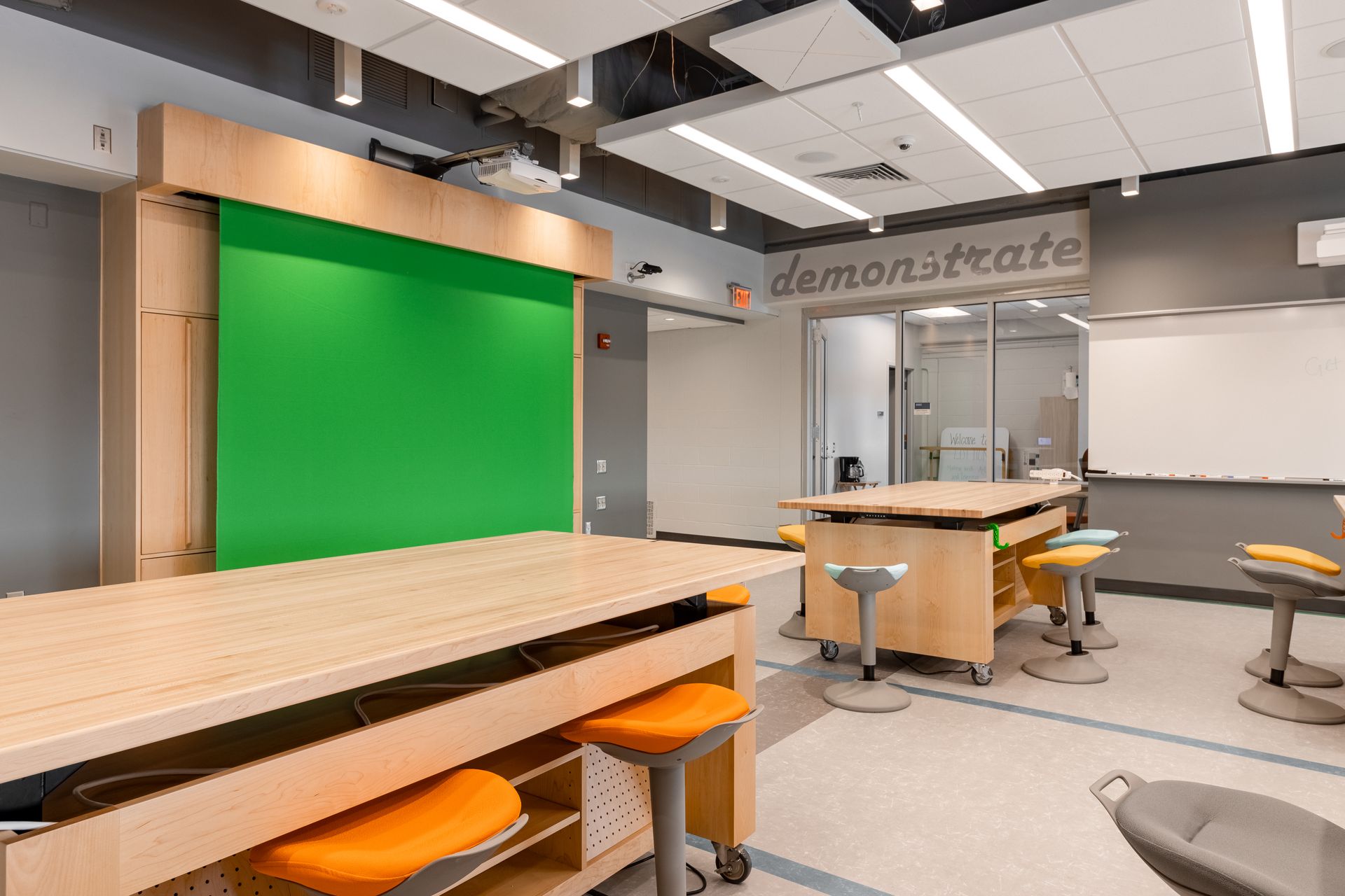 Bright classroom with green screen, light wood tables, orange stools, and the word