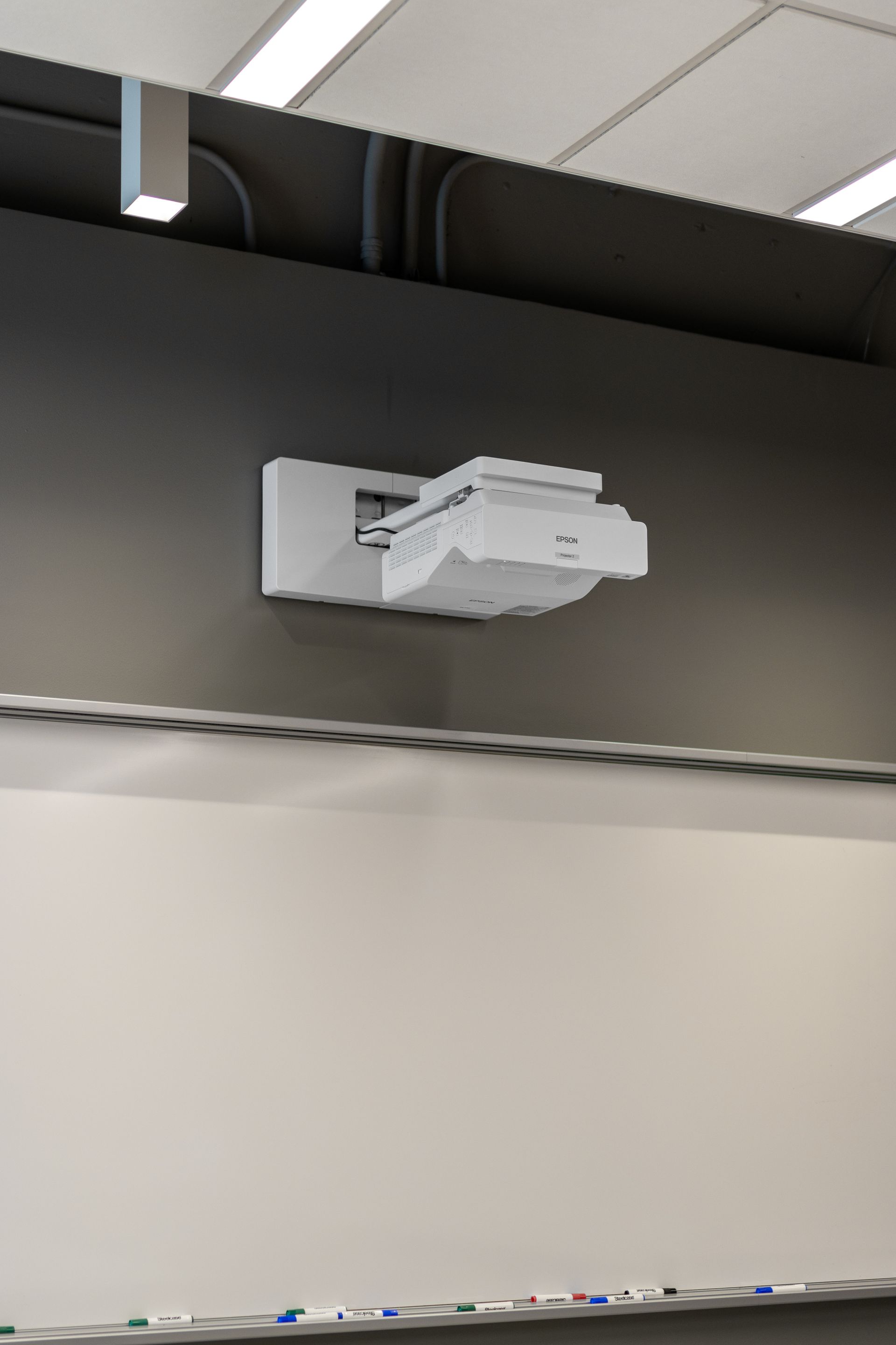 Projector mounted on a dark wall above a whiteboard in a classroom.