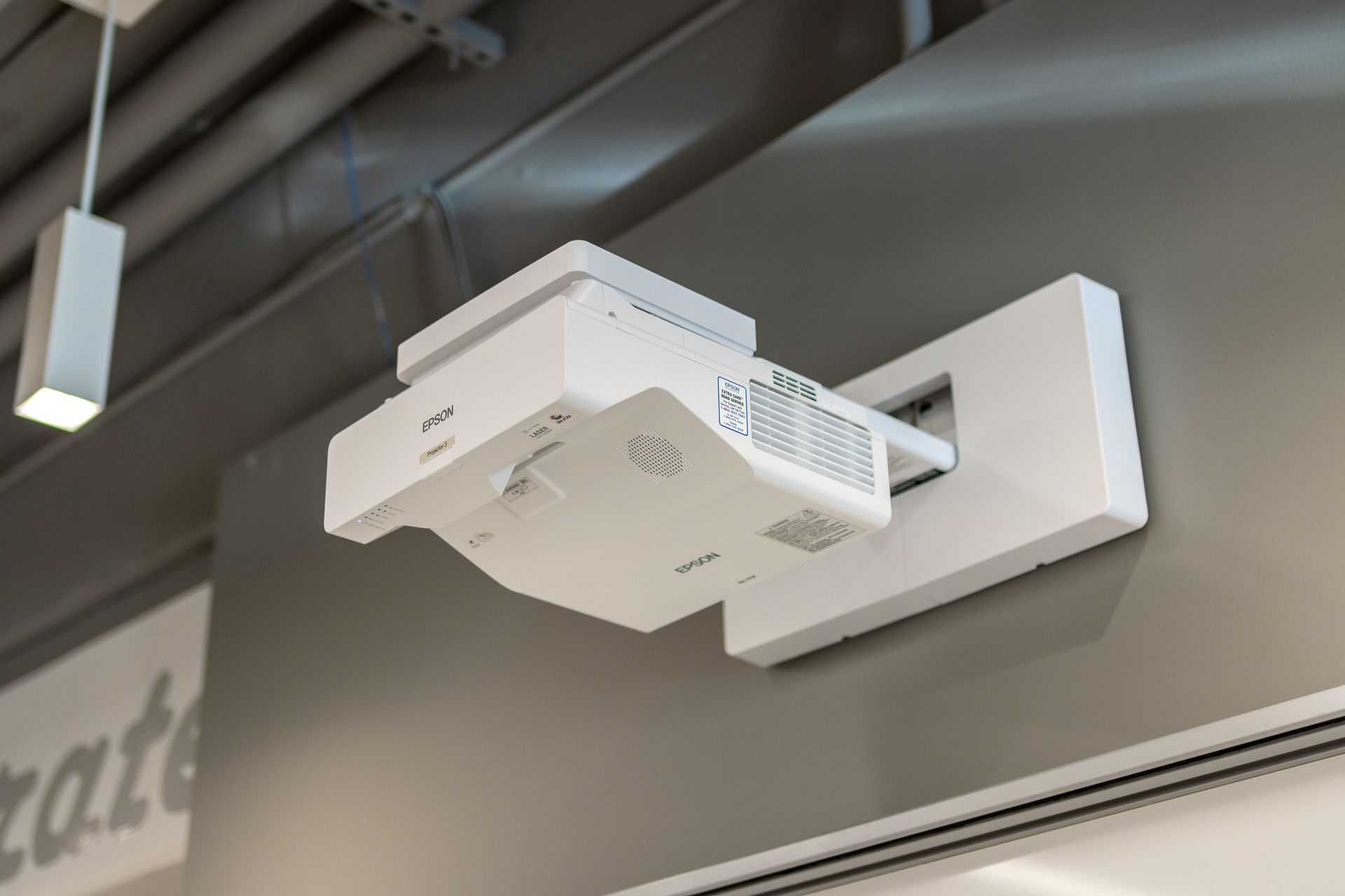 White projector mounted on a white wall bracket. It is attached to a gray wall, near a ceiling with exposed metal piping.