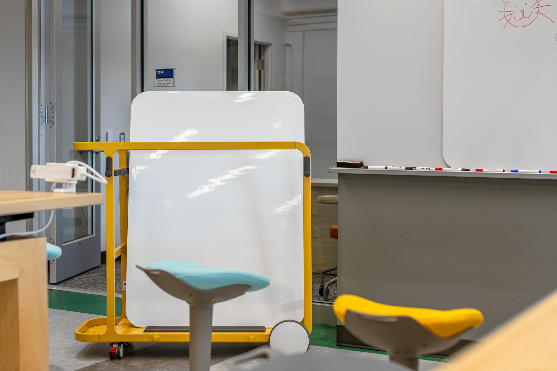 Whiteboard on yellow cart in a room with blue and yellow stools and a partially visible door.
