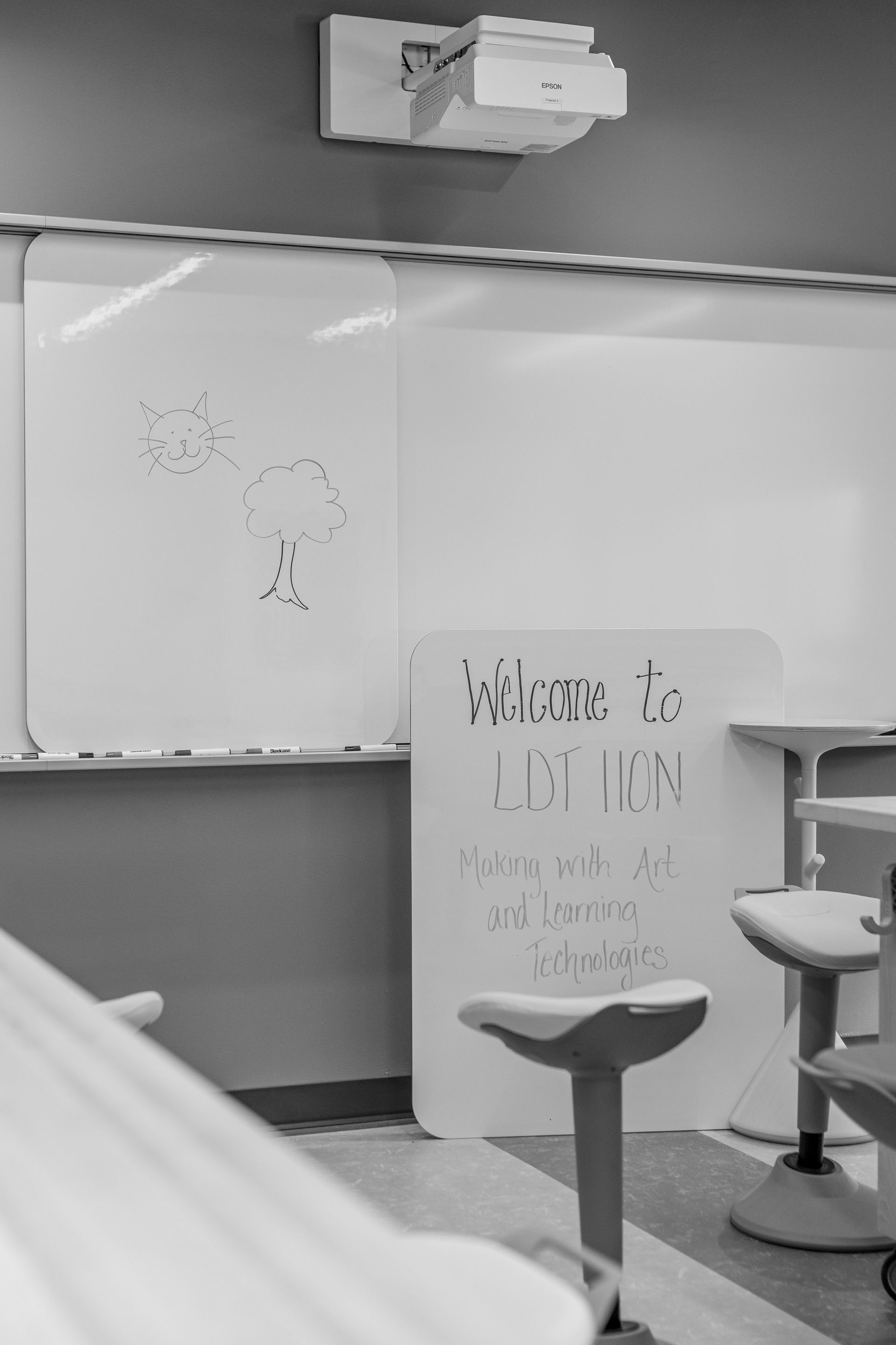 Black and white classroom with whiteboards, a welcome sign, and modern stools. A projector is mounted on the ceiling.