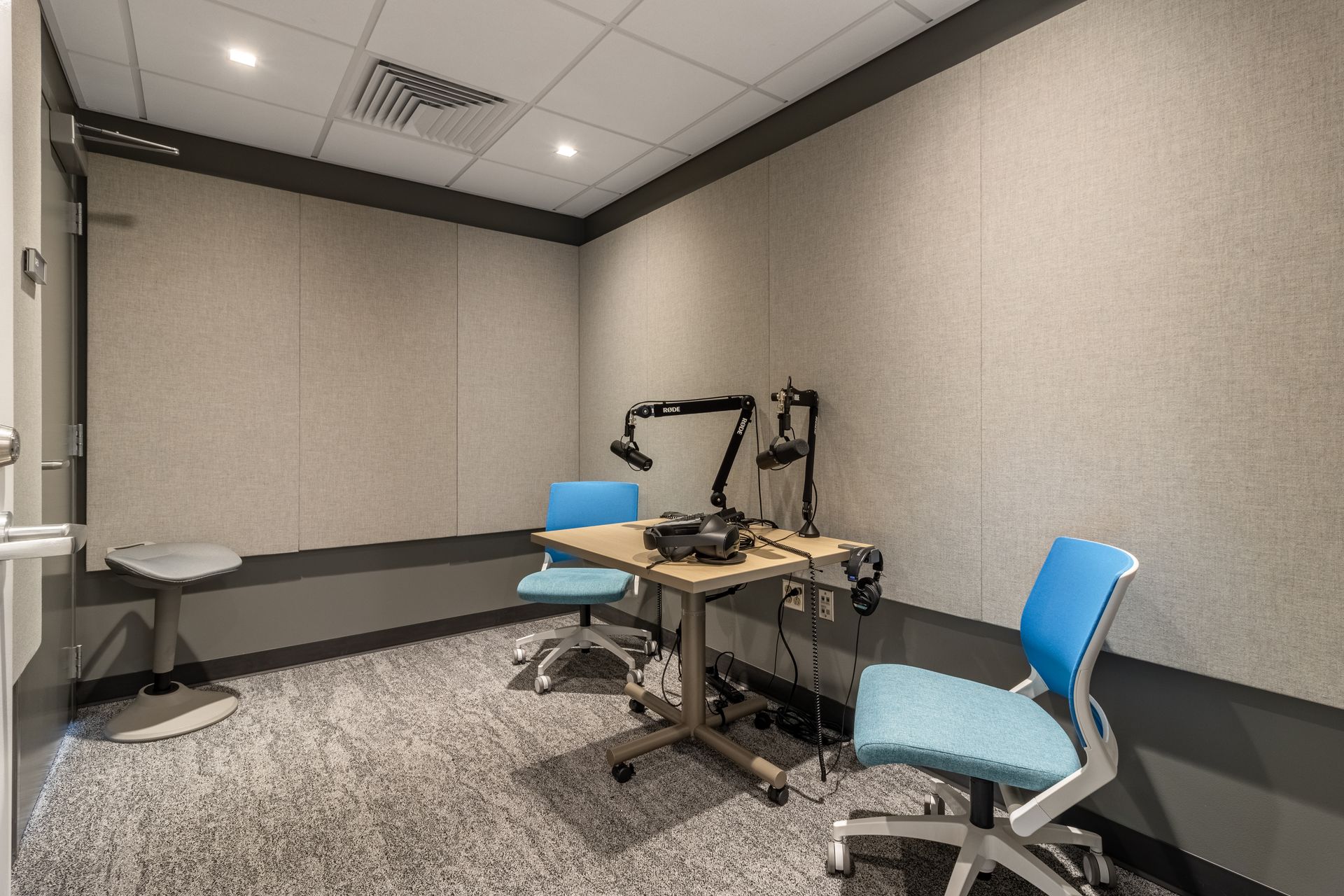 Small soundproof room with two chairs, desk, and equipment. Light blue chairs, gray walls and flooring.