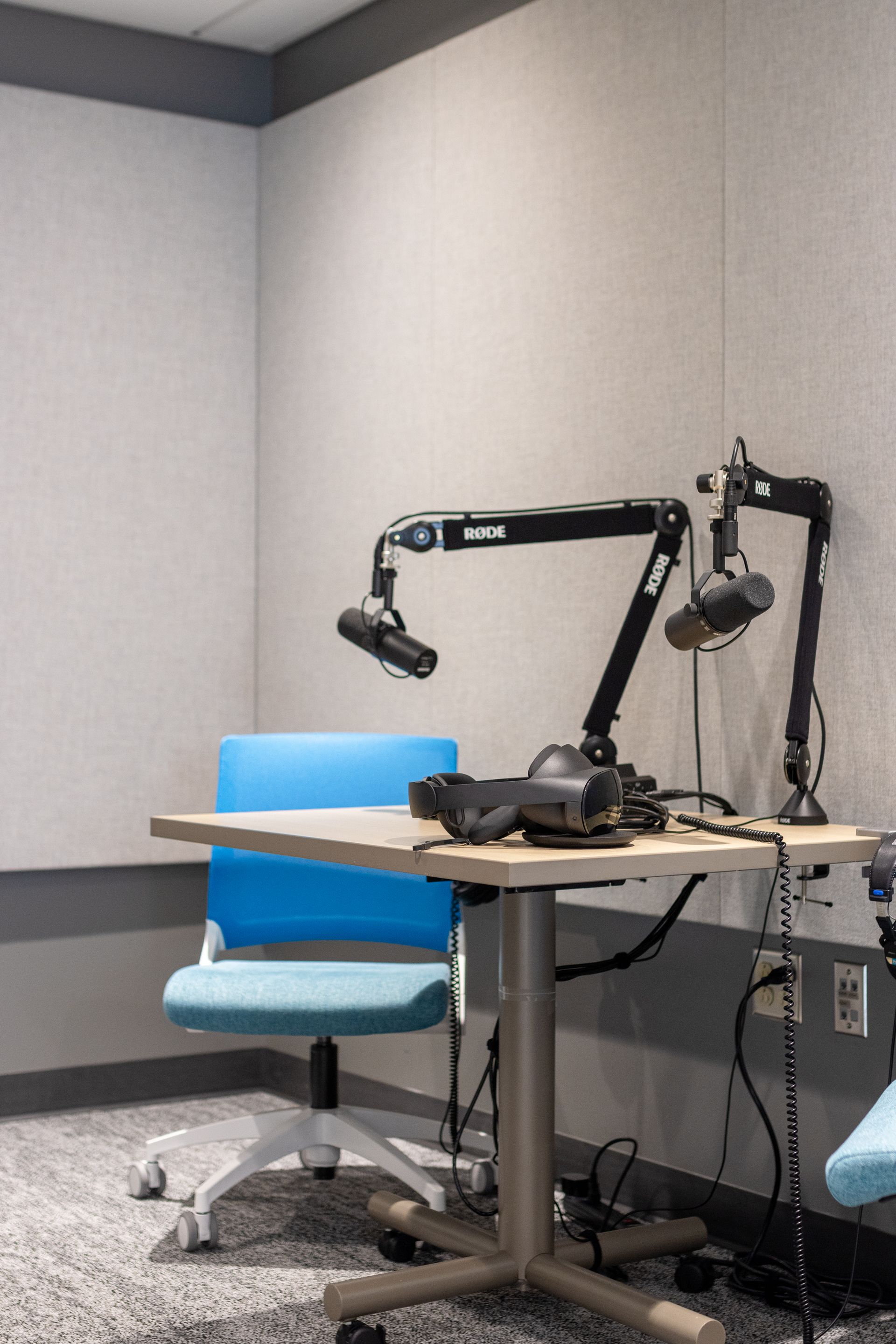 A podcast studio with microphones, table, and blue chair. Gray soundproofing, gray walls, light wooden table, and blue chair.