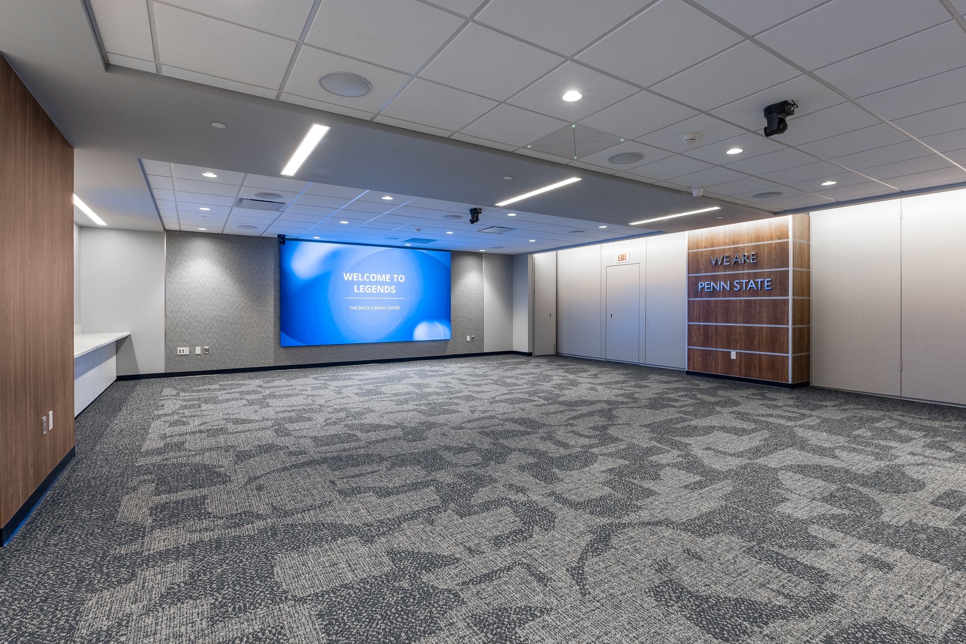Bryce Jordan Center's empty conference room with gray carpet, wood walls, and a blue presentation screen at the front.