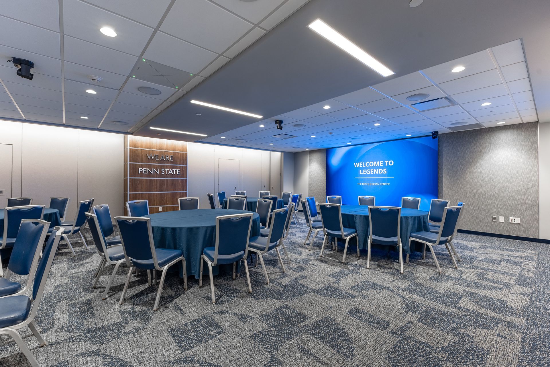 Bryce Jordan Center's empty conference room with round tables, blue chairs, and a blue presentation wall.