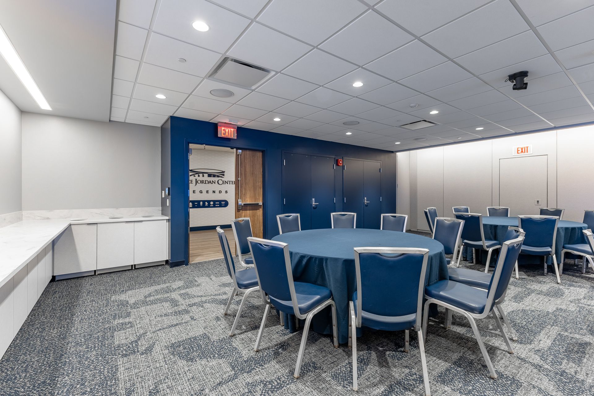 Bryce Jordan Center's empty conference room with blue chairs, round tables, and a carpeted floor near a wooden door.