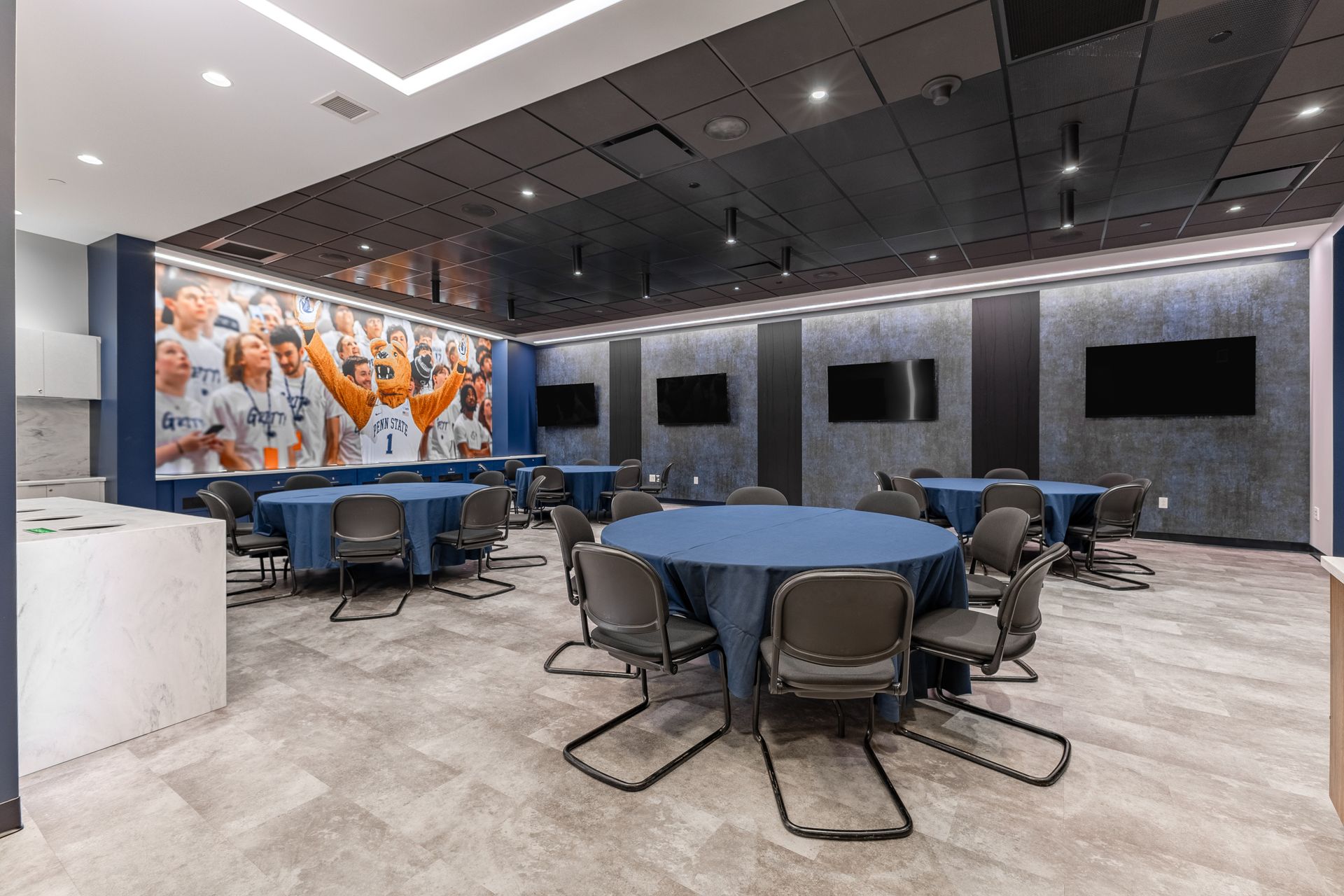Bryce Jordan Center's modern lounge with round tables, chairs, wall-mounted TVs, and a colorful mural on the left.