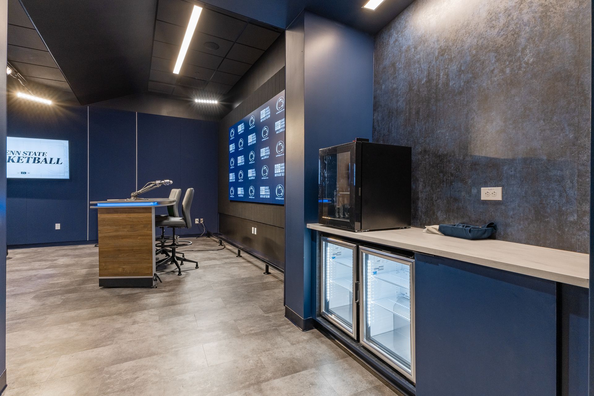 Bryce Jordan Center's modern lounge with blue walls, concrete floor, counter, TV, and chairs in a sleek interior.