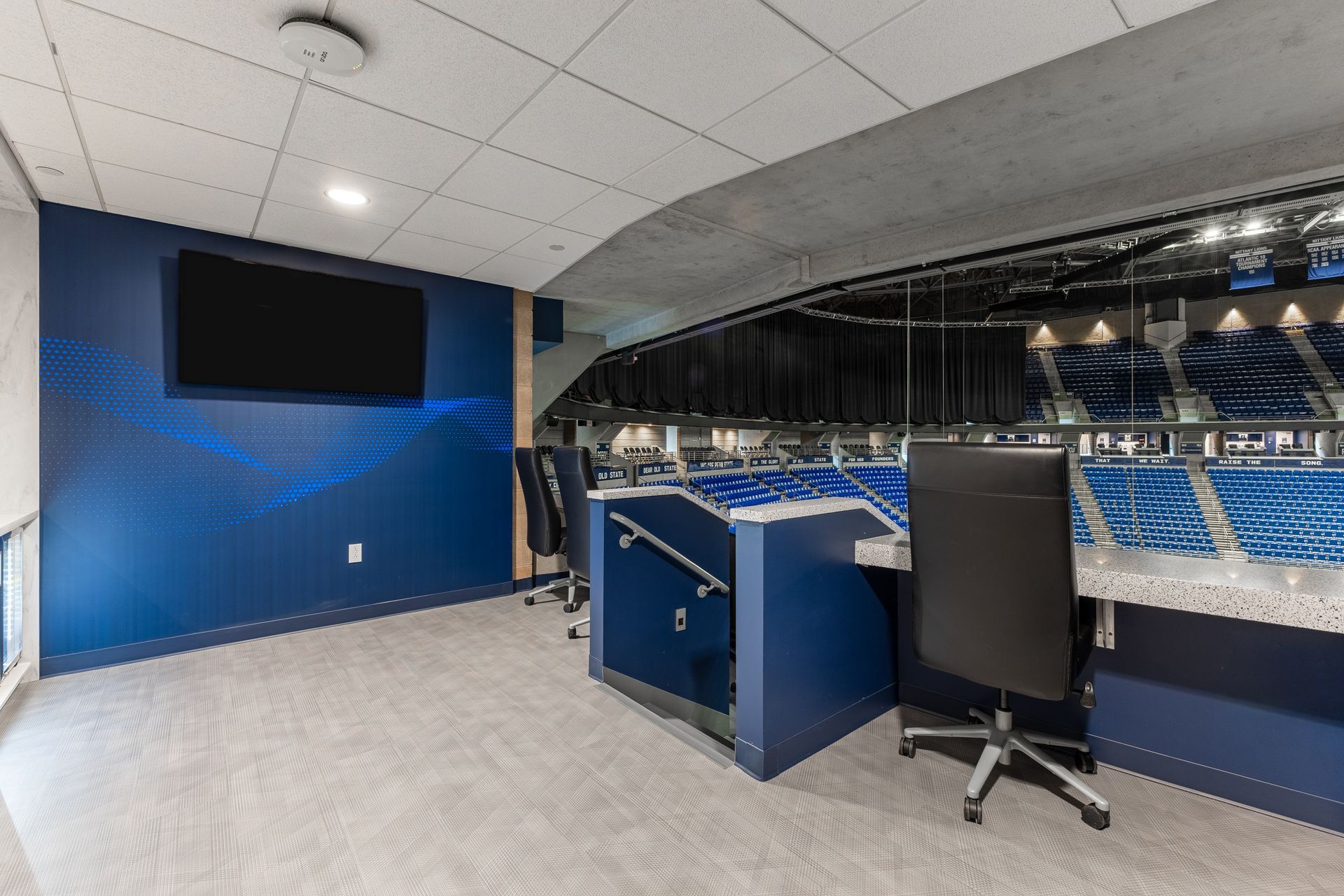 Bryce Jordan Center's empty press box with blue walls, black monitor, desk, and chair overlooking a stadium seating area