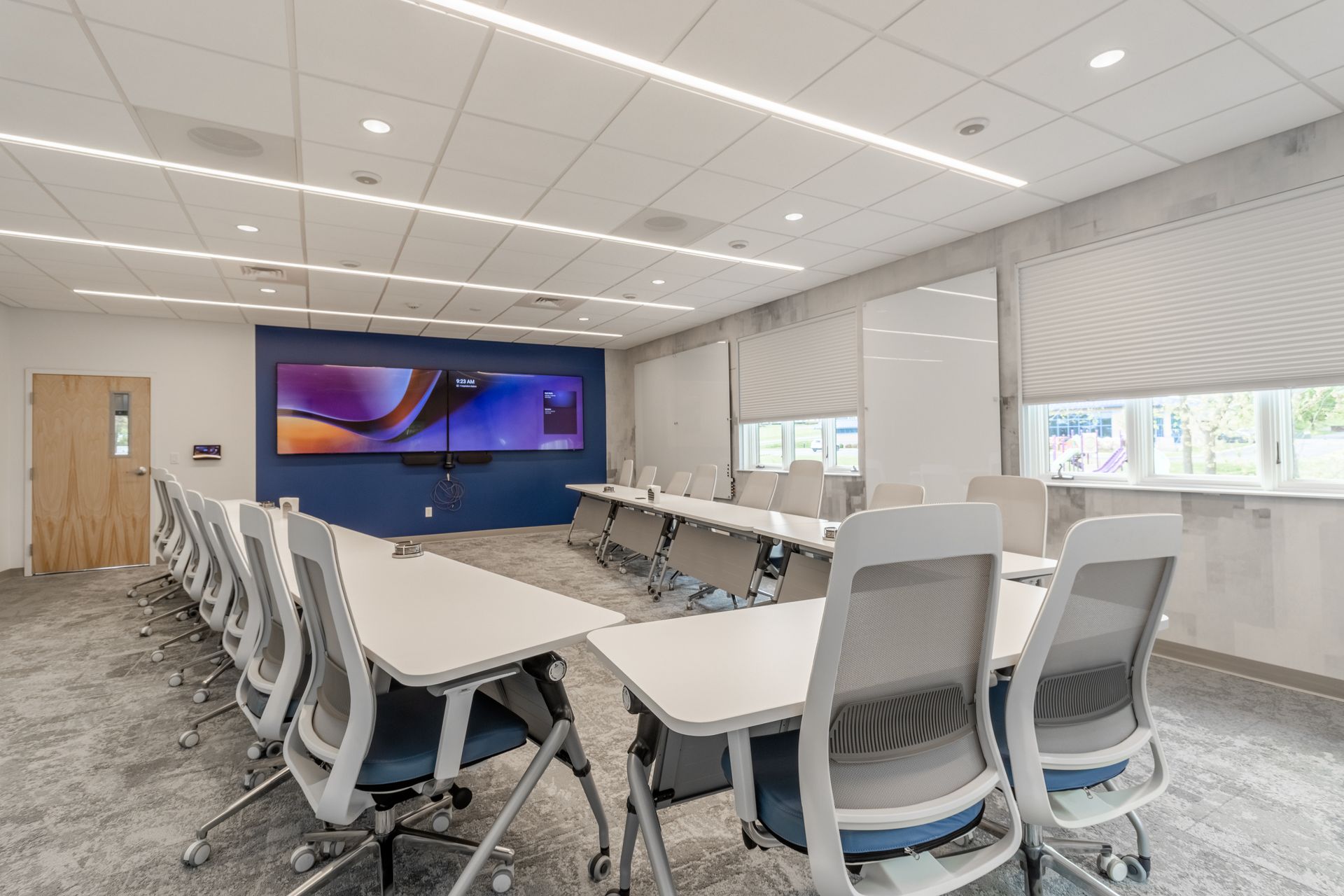 Modern conference room with long tables, gray chairs, wall display, and large windows.