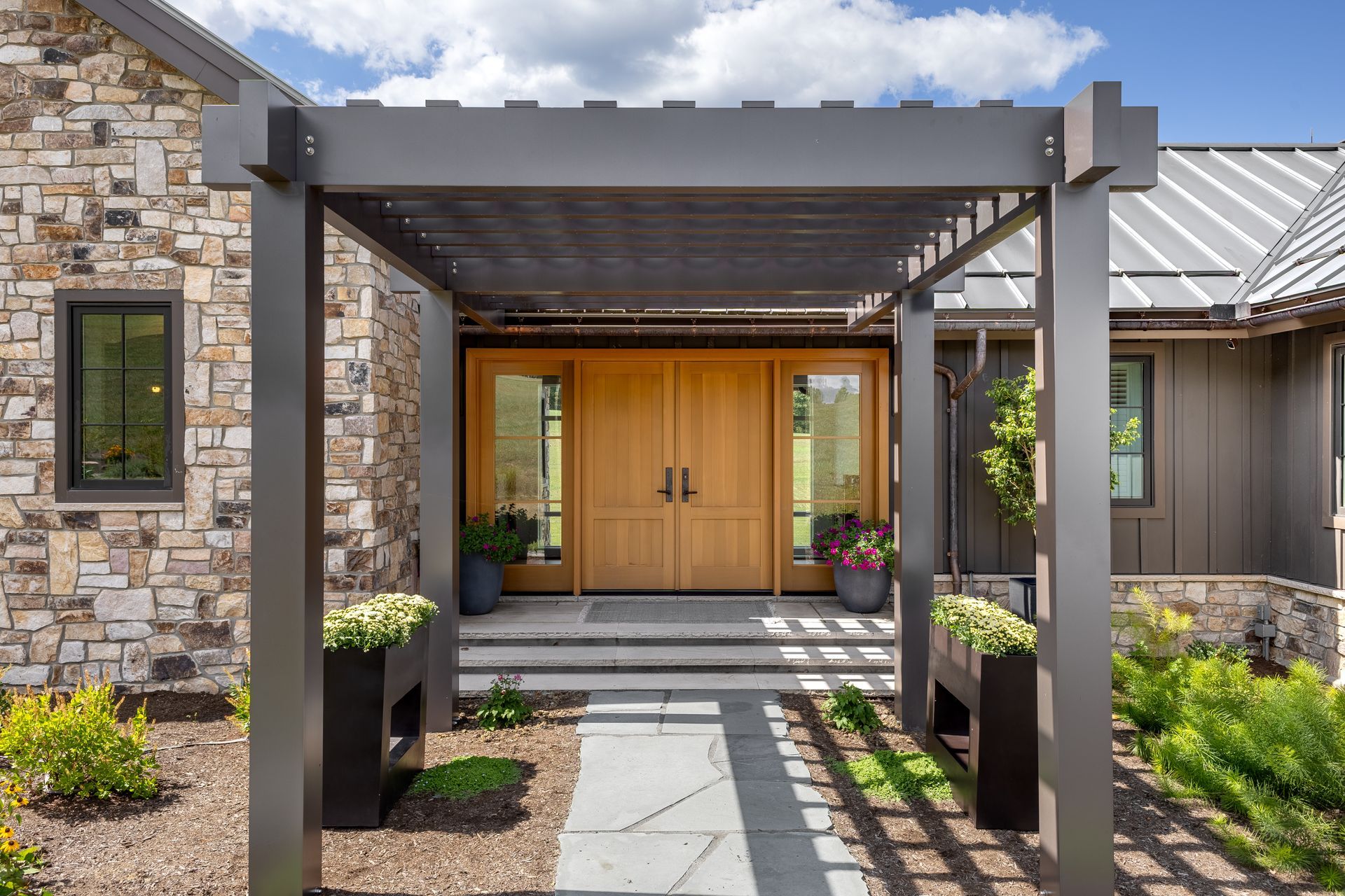 A modern wooden double door entrance framed by a dark grey pergola and stone exterior walls on a sunny day.