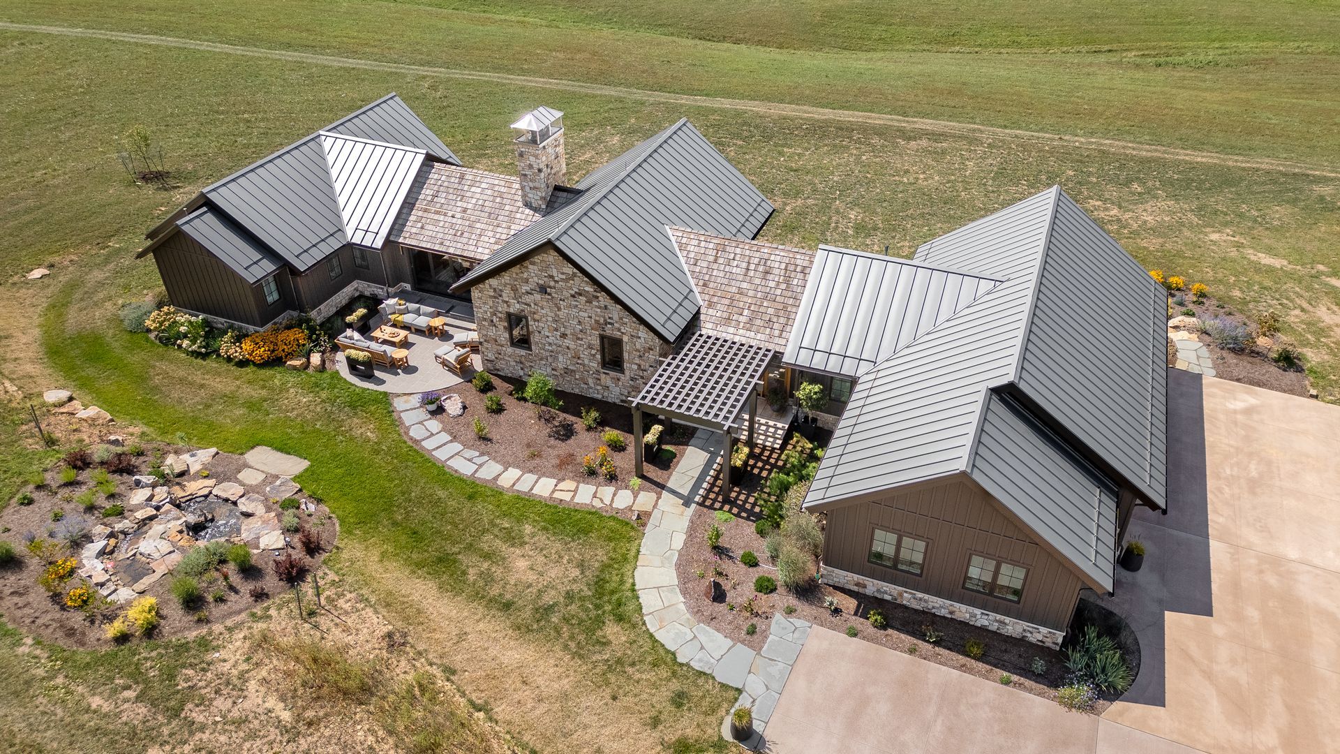 An aerial view of a stone-walled house with a patterned metal roof, surrounded by a rural landscape and stone pathways.