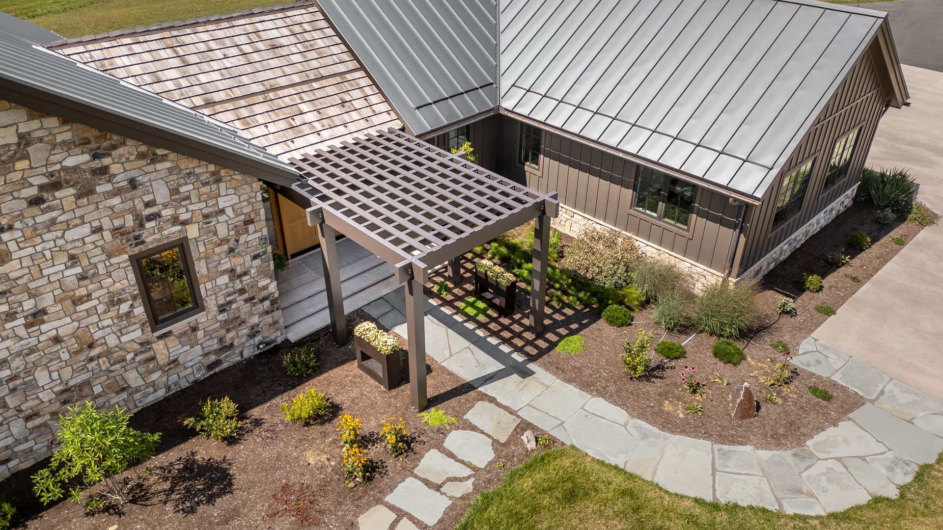 An aerial view of a stone house with a metal roof and a wooden pergola covering a stone walkway leading to the entrance.