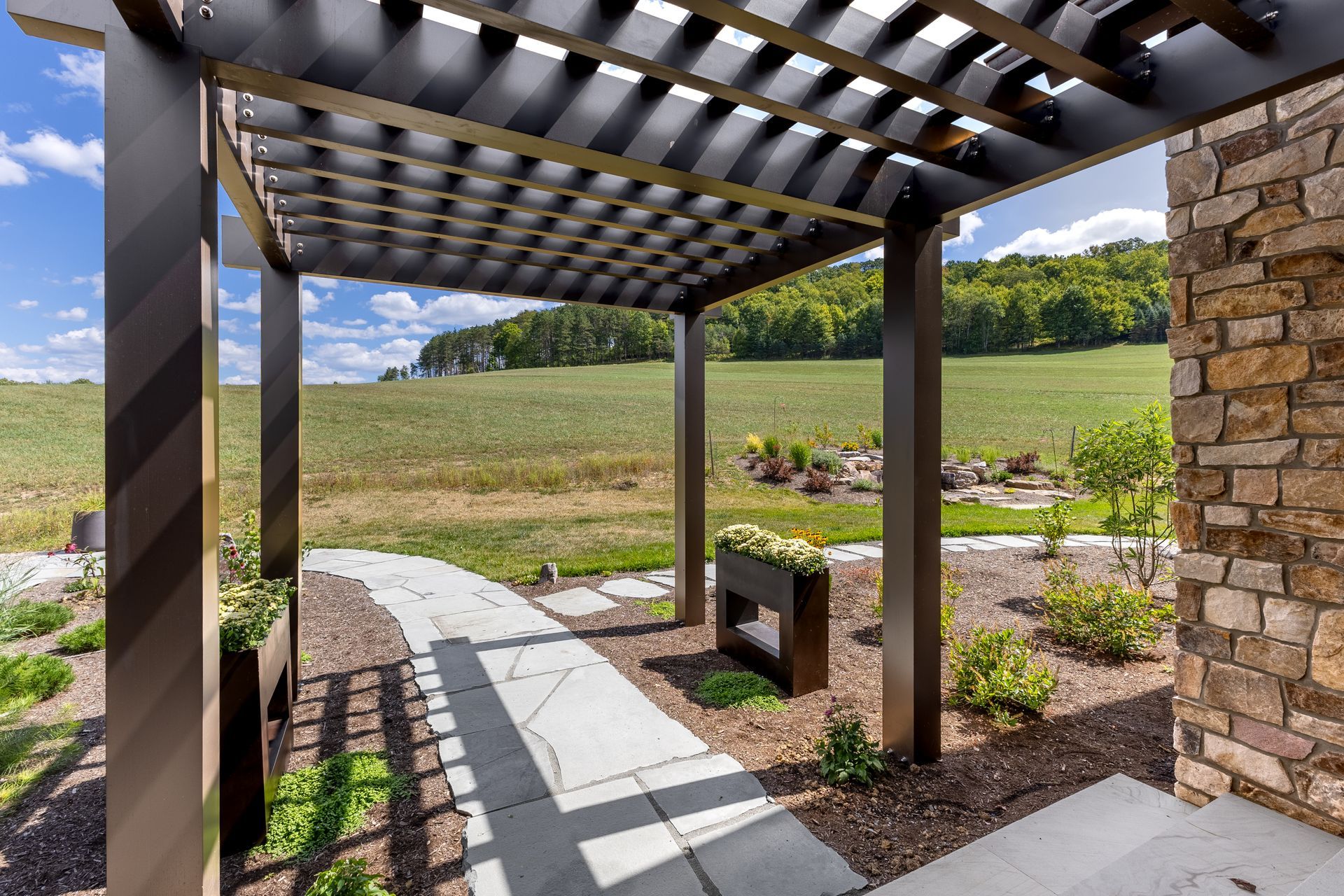 A stone-walled patio with a dark metal pergola overlooking a green, rolling hillside under a partly cloudy blue sky.