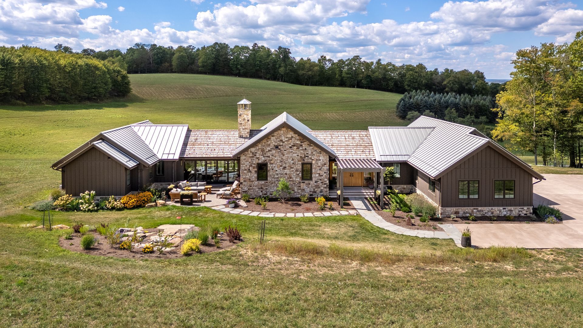 A large, single-story house with stone and dark wood siding, a metal roof, and a grassy lawn in a rolling rural landscape.