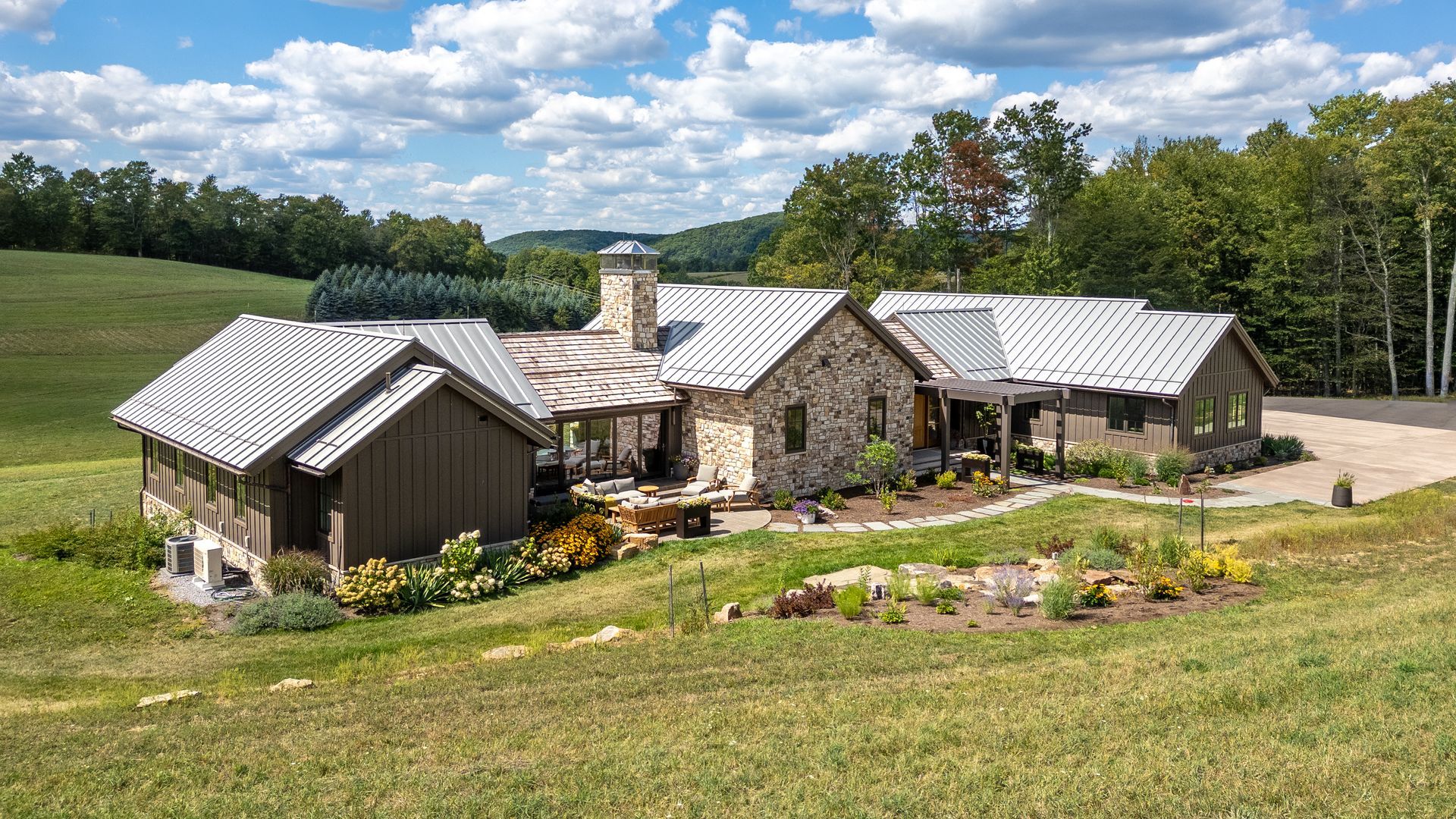 A stone house with a metal roof surrounded by lush green fields and trees under a cloudy blue sky.