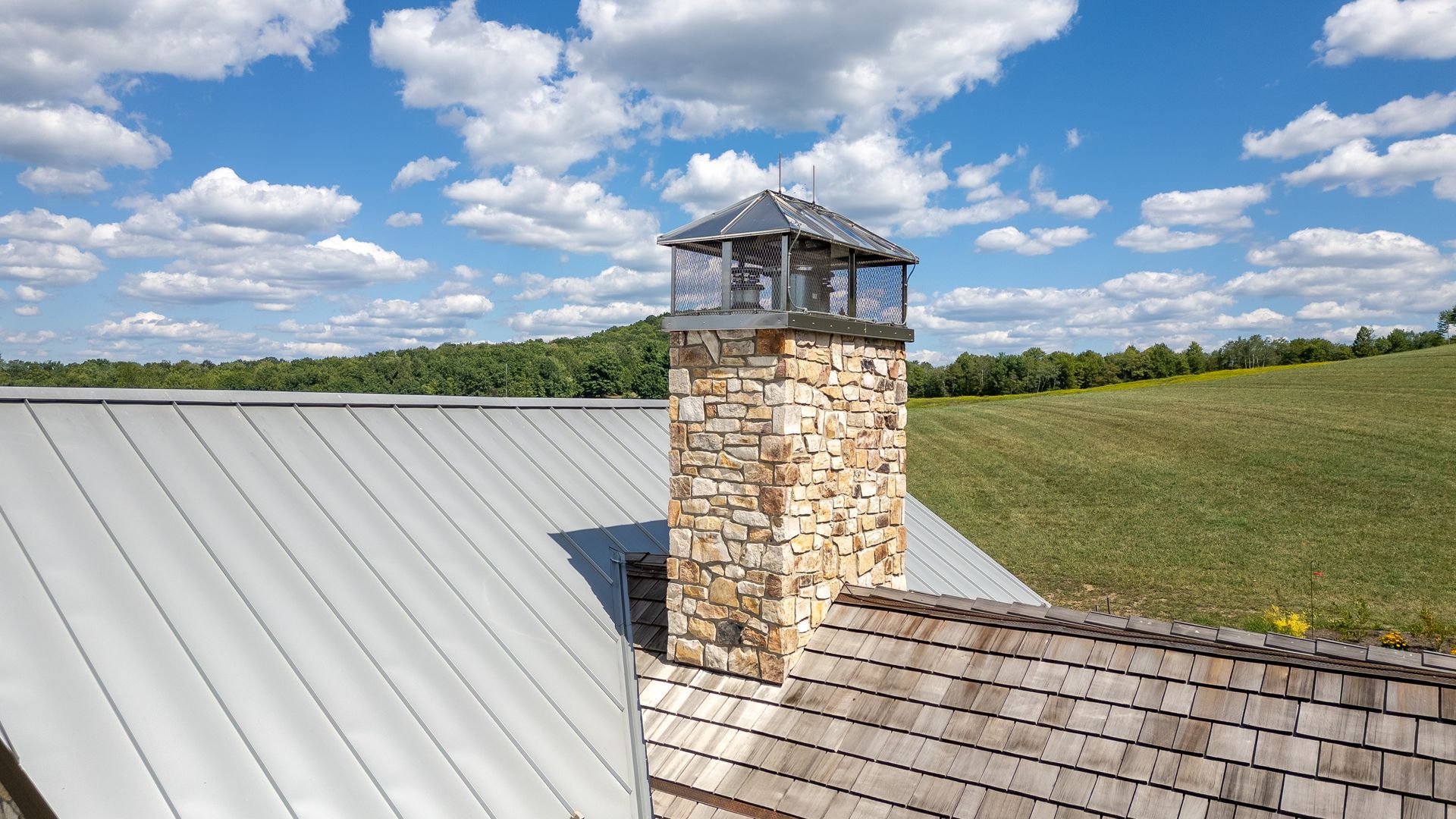A stone chimney with a metal cap sits between a gray metal roof and a wooden shingle roof under a blue, cloudy sky.
