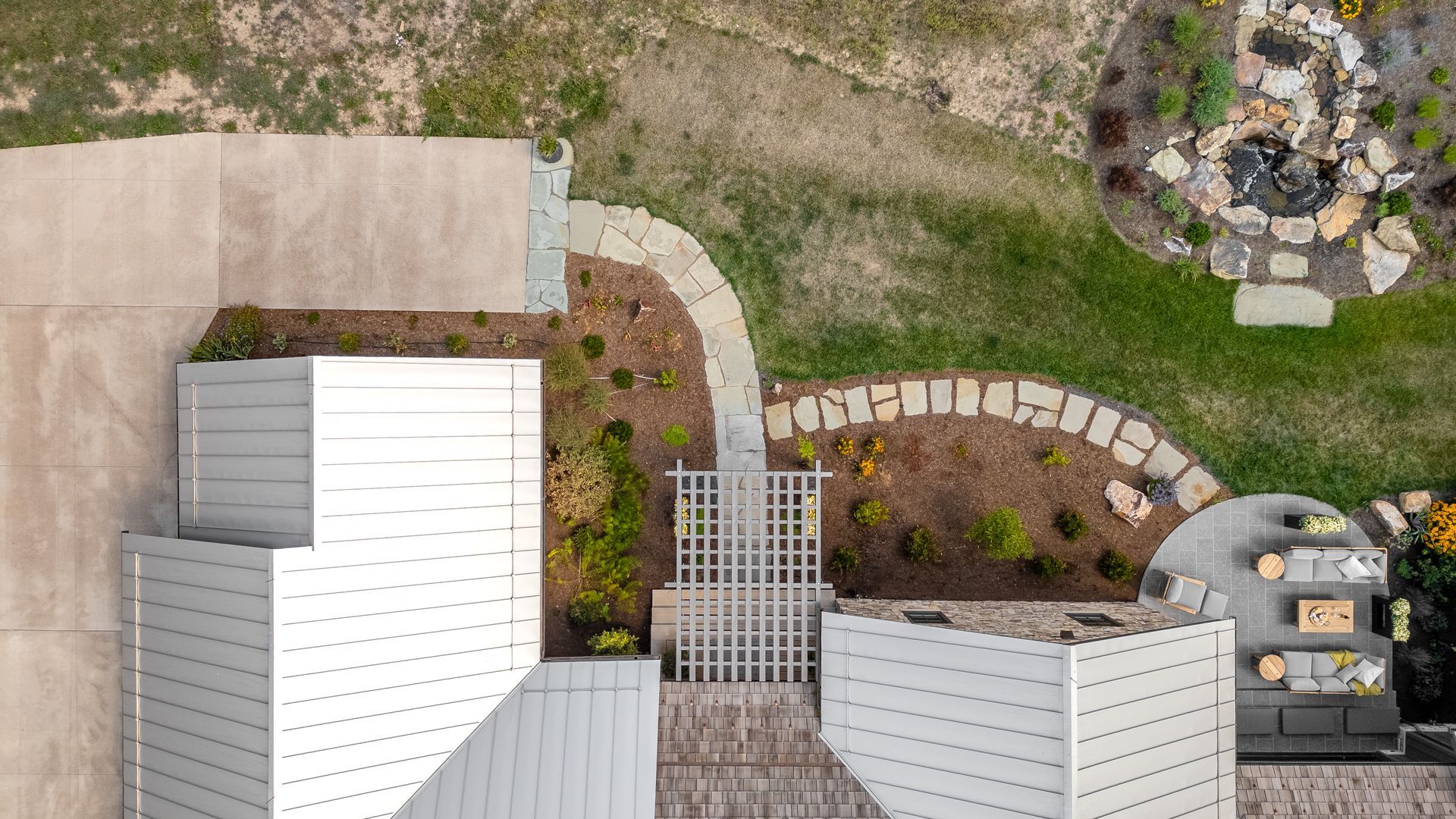 An aerial view of a house roof, a pergola, a stone walkway, landscaping, and a patio with chairs in a grassy backyard.