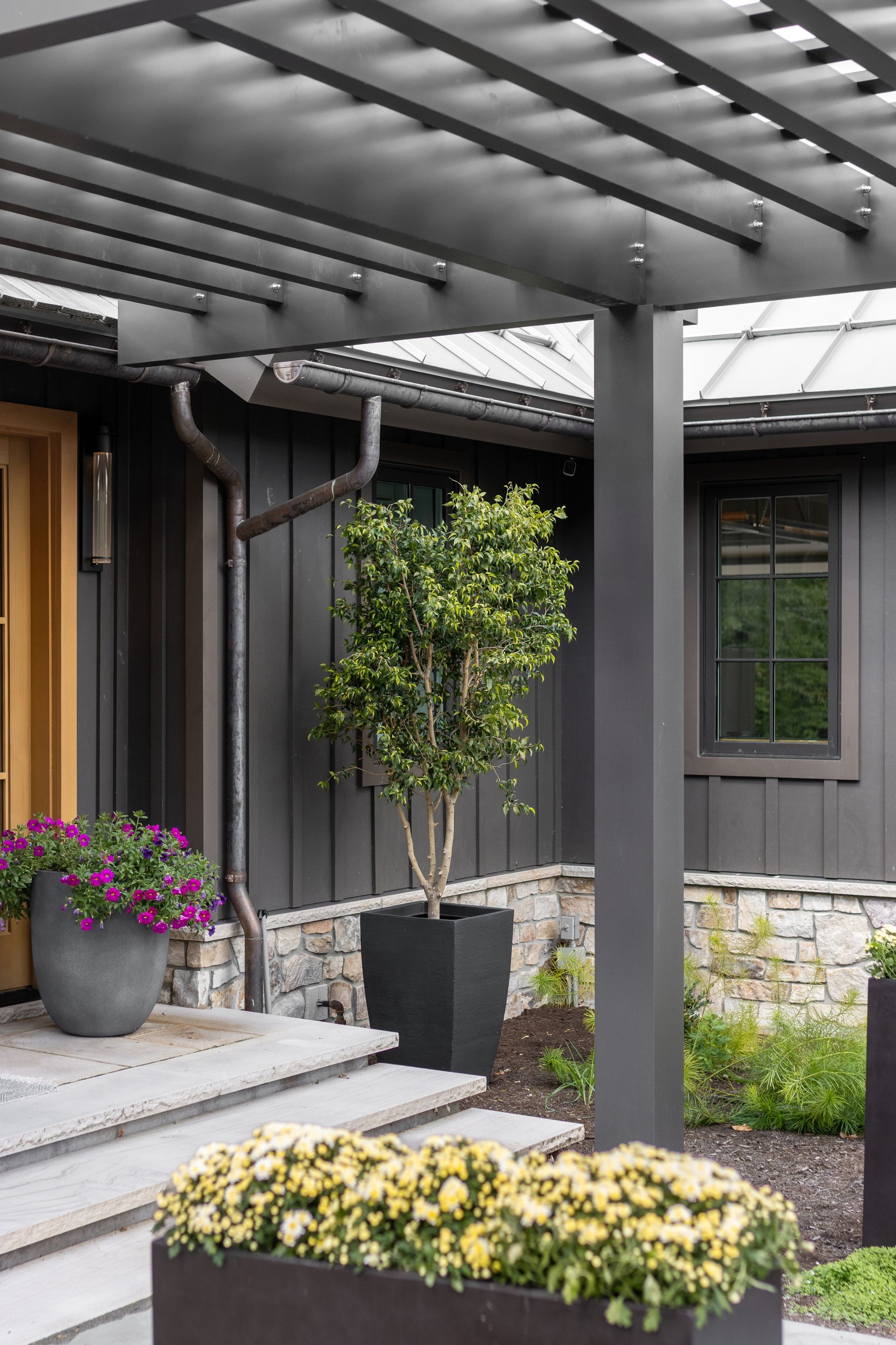 A modern home entrance with a dark wood pergola, steps, a stone foundation, and planters filled with flowers and greenery.