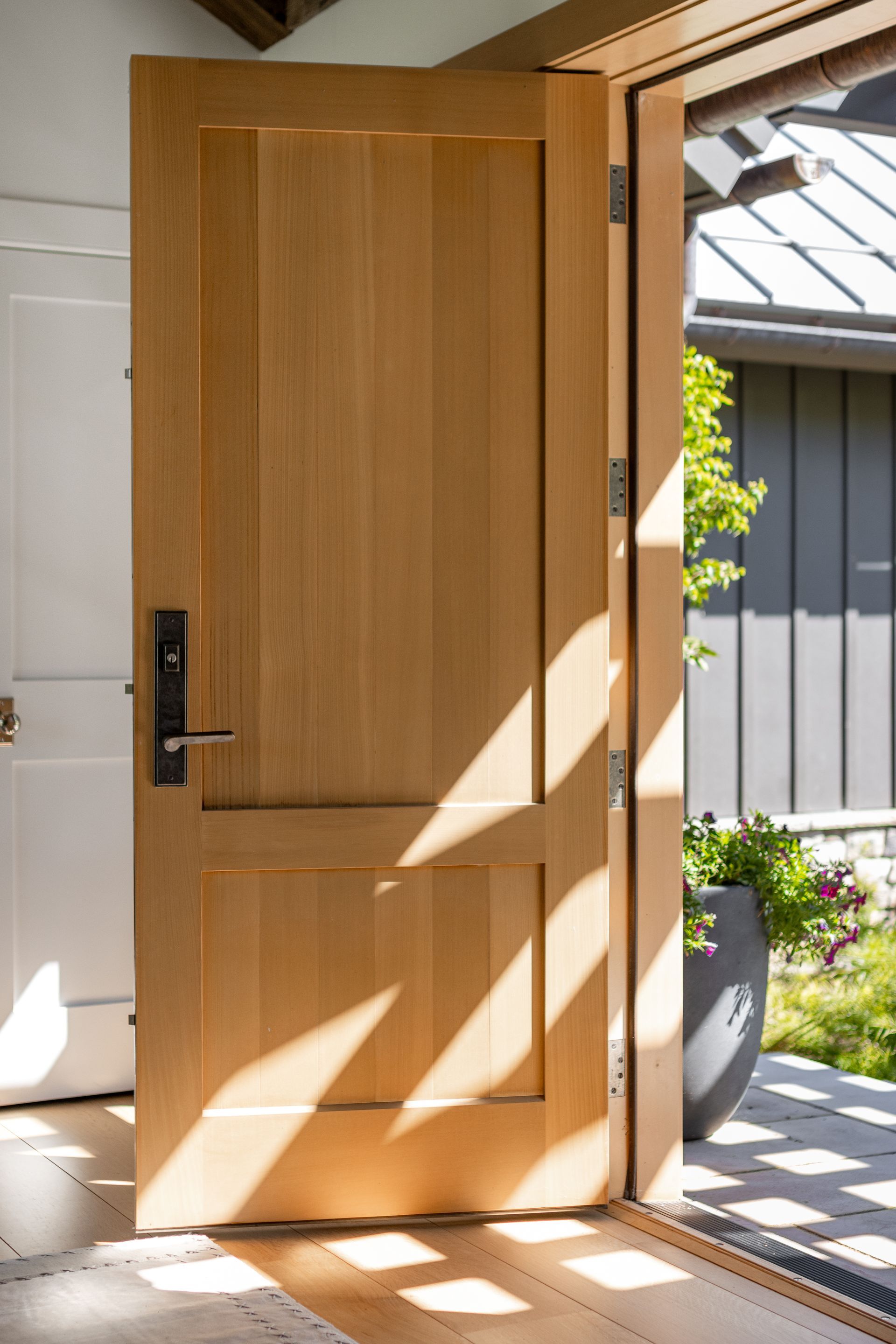 A light wood door stands slightly ajar, casting long, geometric shadows across a bright, sunlit indoor entryway.