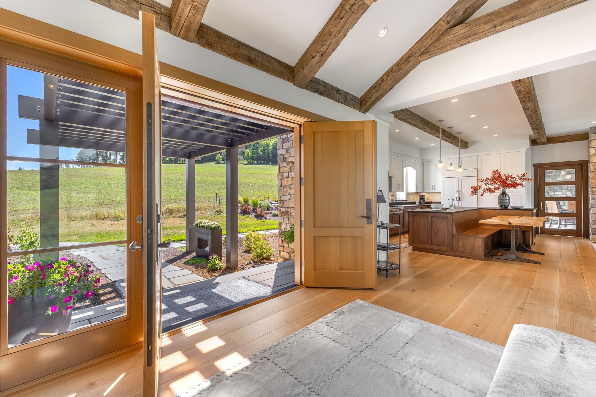 A wide-open wooden door leads to a patio with a pergola, revealing a grassy hill beyond a bright, modern interior room.