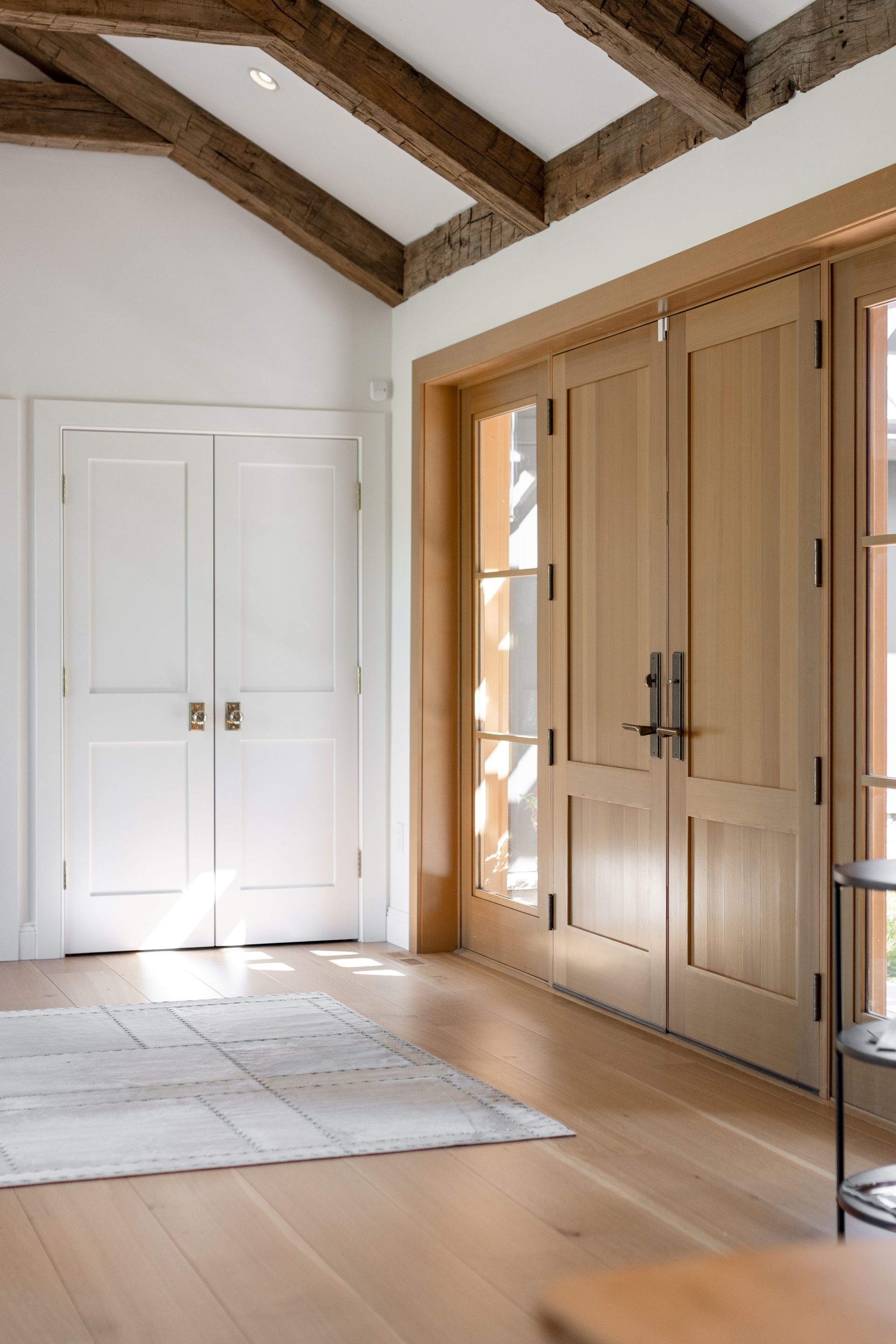 An entryway with light wood floors, white walls, exposed ceiling beams, a white double door, and tall glass-paneled doors.
