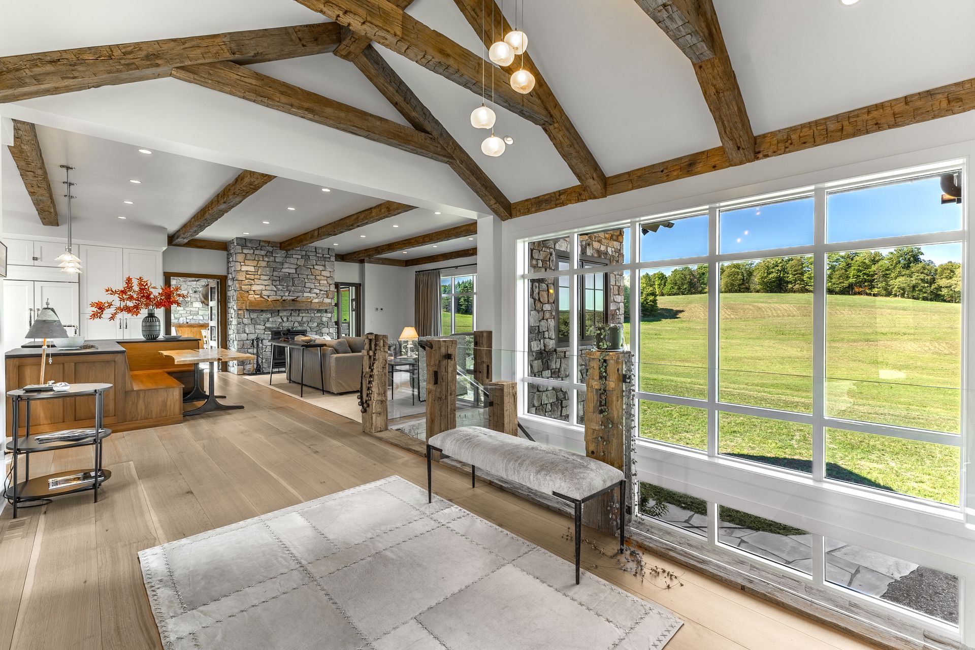 A light-filled open-plan room with wood-beamed ceilings, stone accents, and a large window wall overlooking a green field.