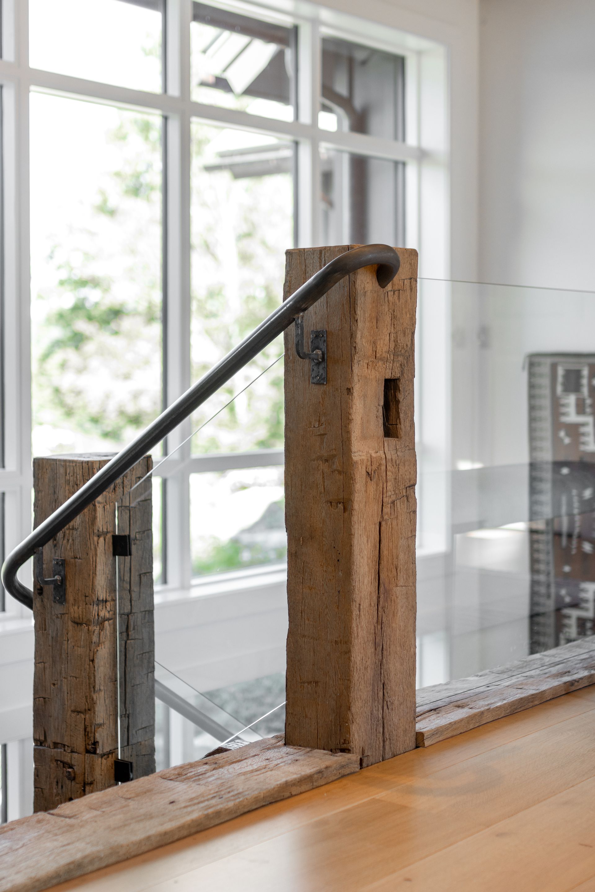 Rustic wooden posts supporting a thin metal handrail and glass panels against a windowed background.