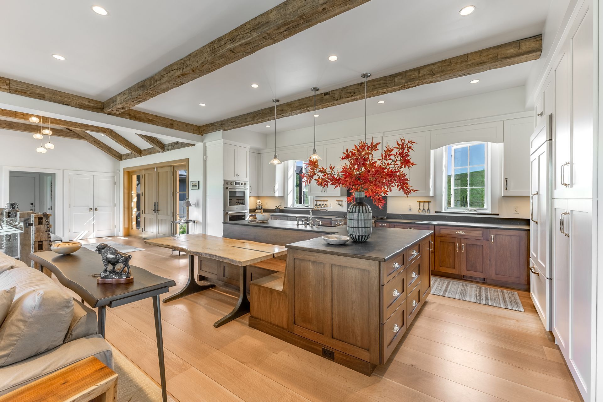 A bright, open-concept kitchen with rustic wood ceiling beams, a central wood island, a dining table, and white cabinetry.