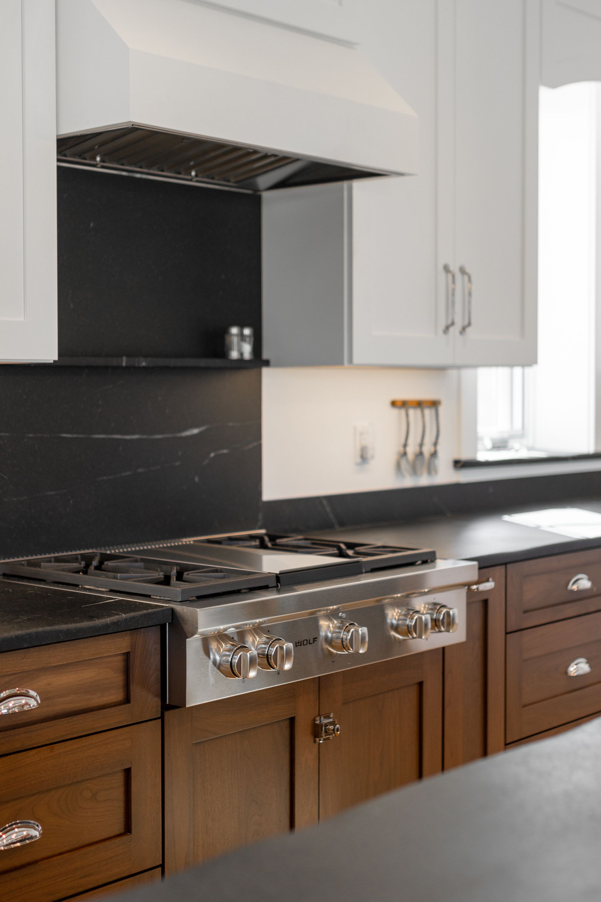 A kitchen stovetop with a stainless steel range, dark stone backsplash, and wooden cabinets below white upper cabinets.